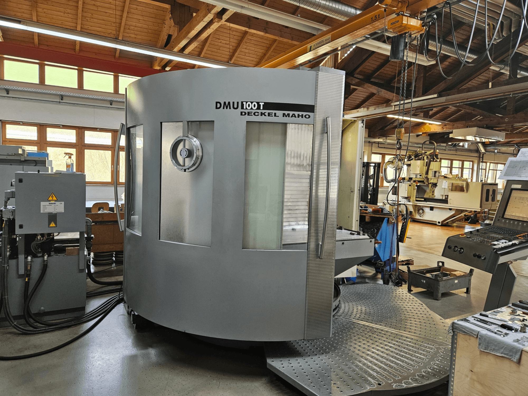 DMU 100 T Deckel Maho CNC-kone edestä katsottuna, jossa on suuri pyöreä pohja, ohjauspaneeli ja lasikotelo.