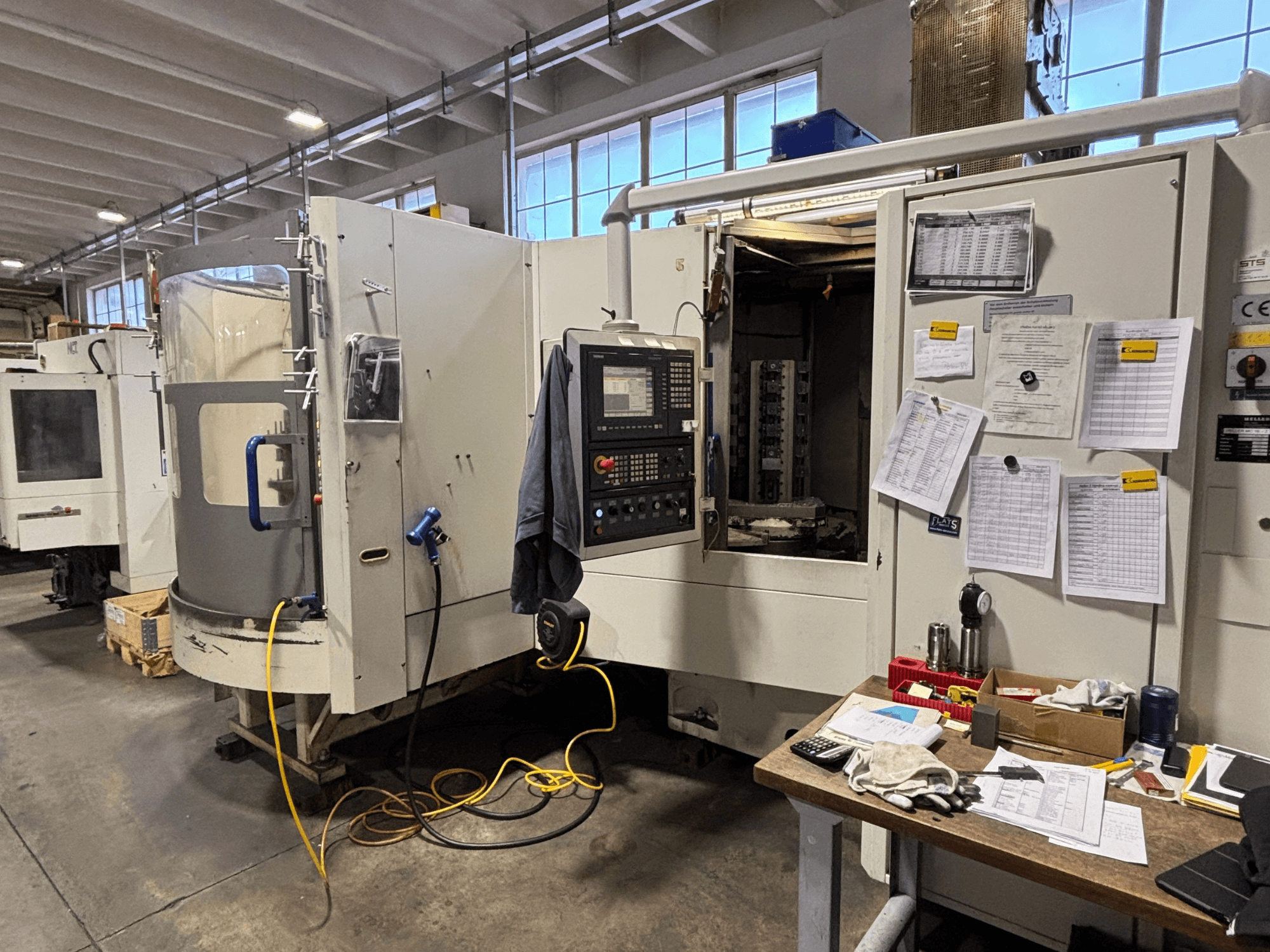 CNC-kone työpajassa, edestä katsottuna, ohjauspaneeli ja dokumentaatio sivulla; työkalut ja materiaalit pöydällä.