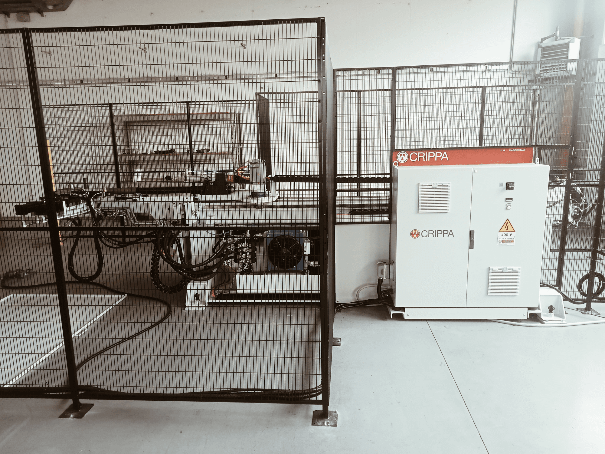 Valkoinen CRIPPA-kone turvahäkin takana, letkut ja komponentit näkyvissä, sivukuva, teollisuusympäristö.