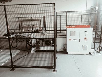 Valkoinen CRIPPA-kone turvahäkin takana, letkut ja komponentit näkyvissä, sivukuva, teollisuusympäristö.