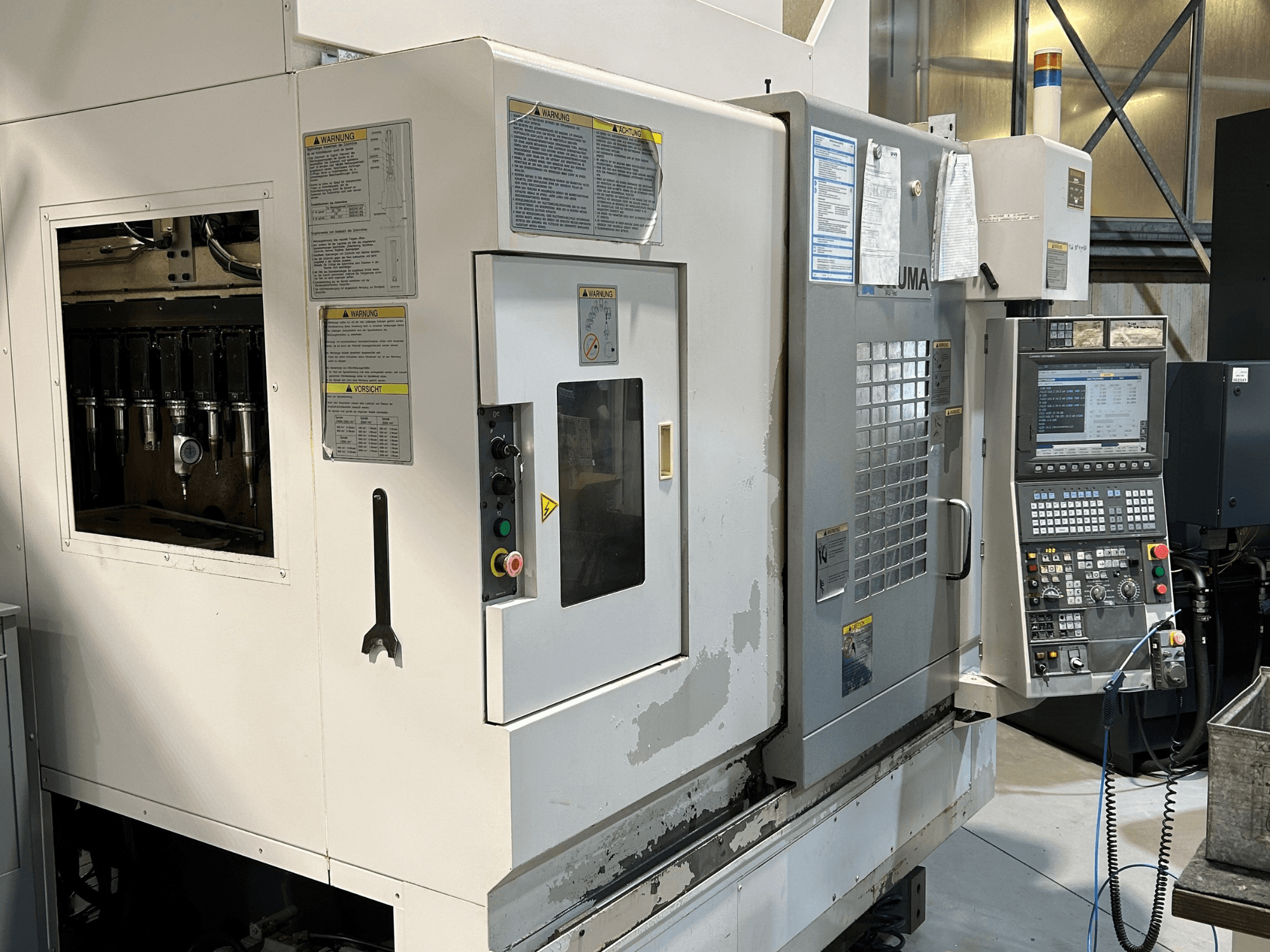 UMA CNC-koneen etukuva, jossa näkyvät työkalunpitimet, ohjauspaneeli, varoitustarrat ja käyttöpainikkeet.