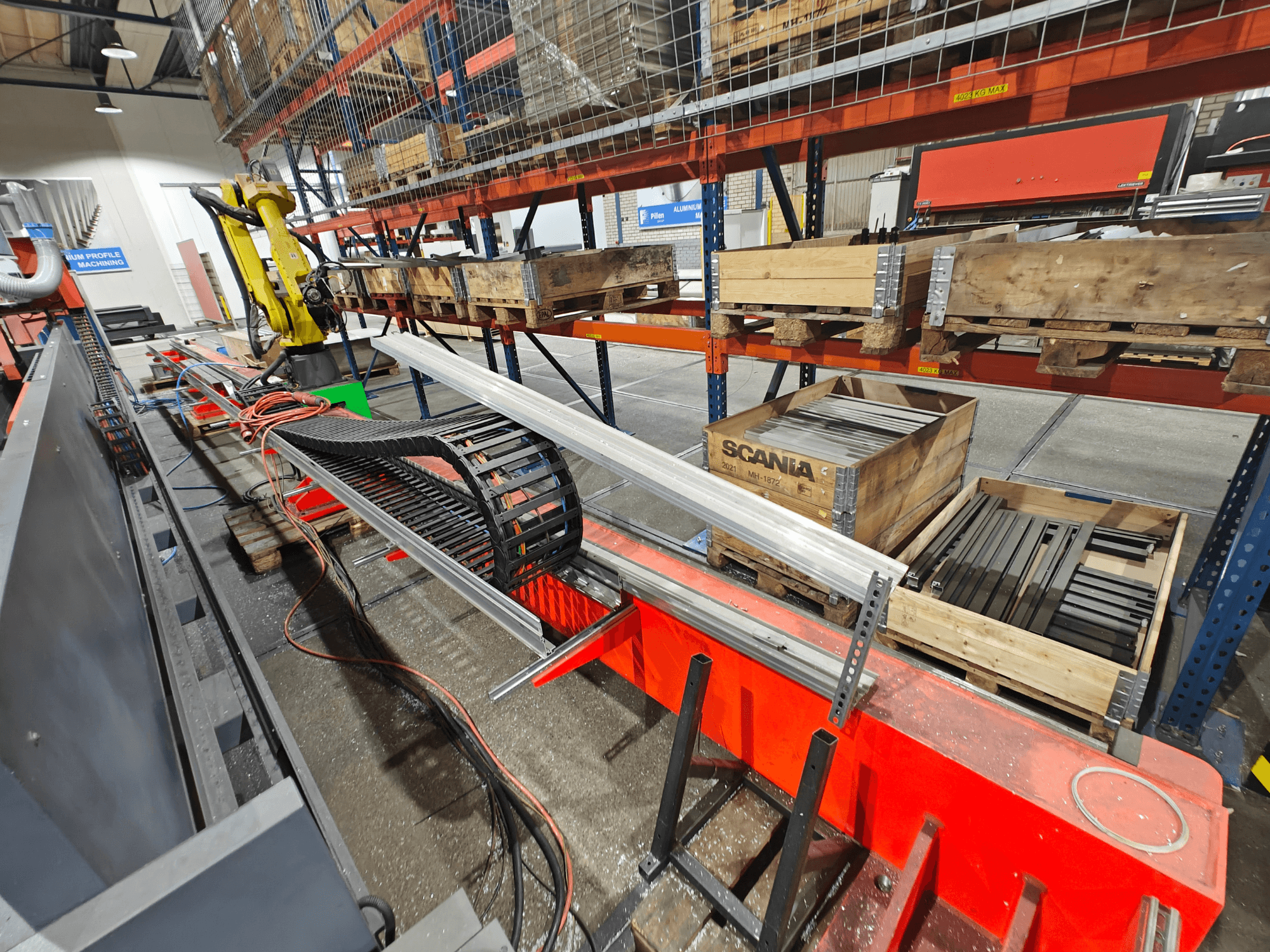 Robottivarsi työpisteessä, oranssit ja mustat kaapelit näkyvissä, Scania-laatikoita ja alumiiniprofiileja tehdasympäristössä.