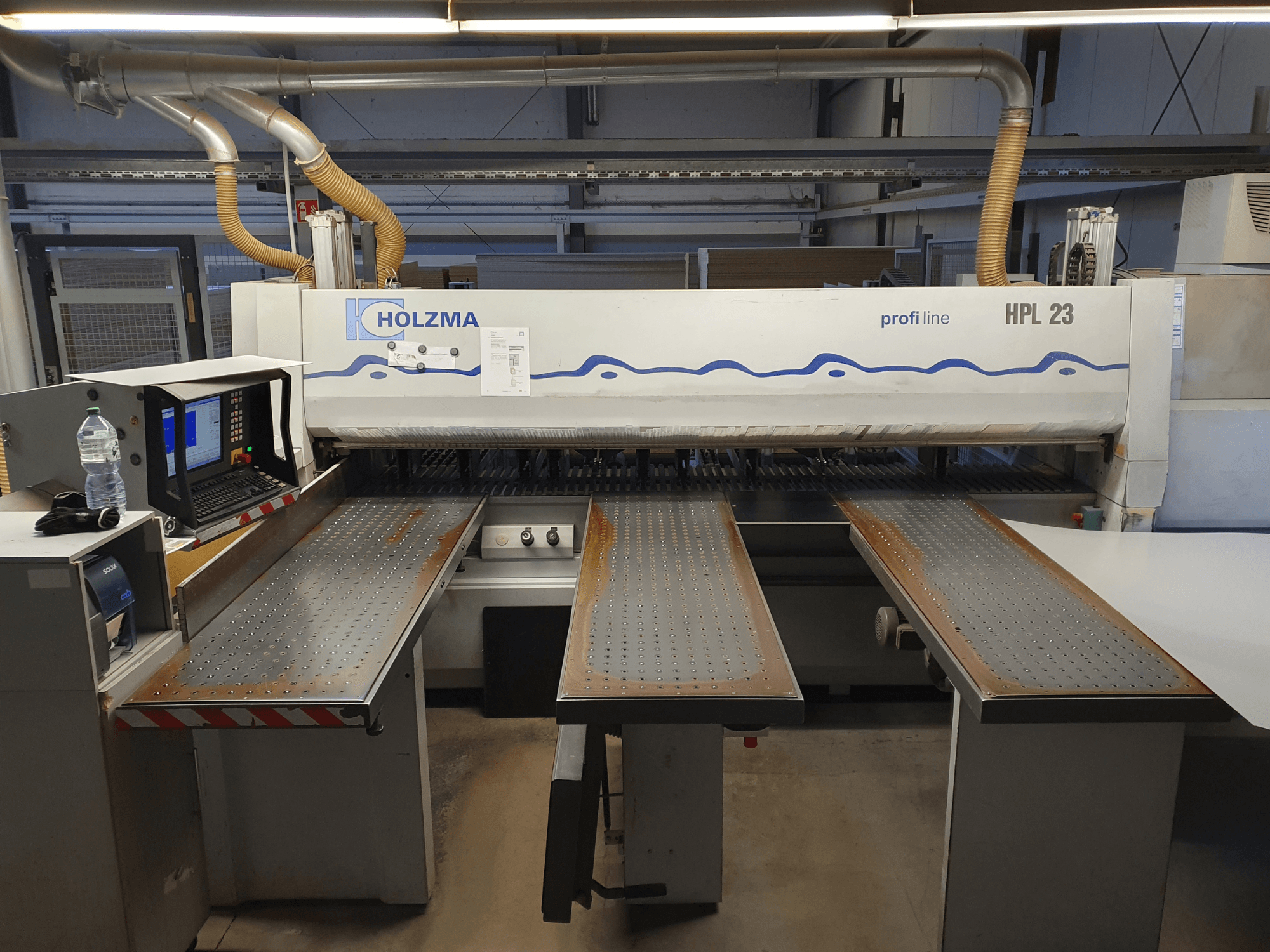 HOLZMA HPL 23 -leikkauskone edestäpäin, kolme rei'itettyä pöytää, tietokonepääte ja vesipullo vieressä.