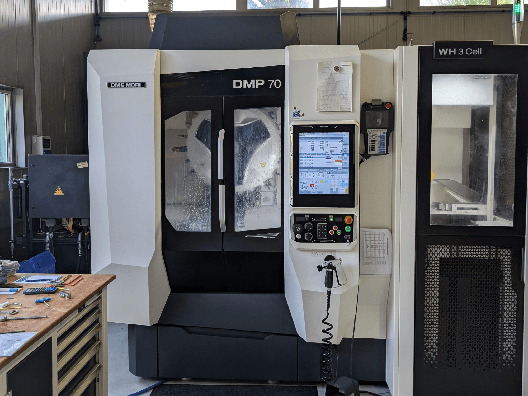 DMG MORI DMP 70 -pystykoneistuskeskus, etunäkymä ohjauspaneelilla ja käyttönäytöllä, työpajan työkalujen ympäröimänä.