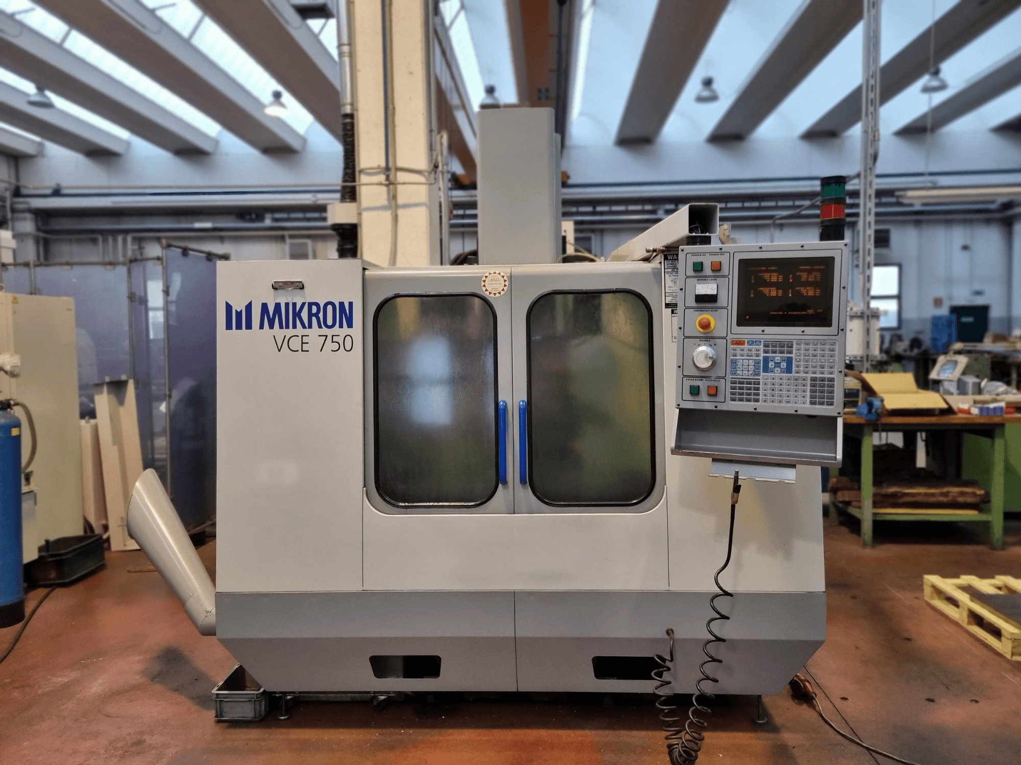 Valkoinen Mikron VCE 750 CNC-kone työpajassa, sivukuva, jossa on digitaalinen ohjauspaneeli ja suuret lasiovet.