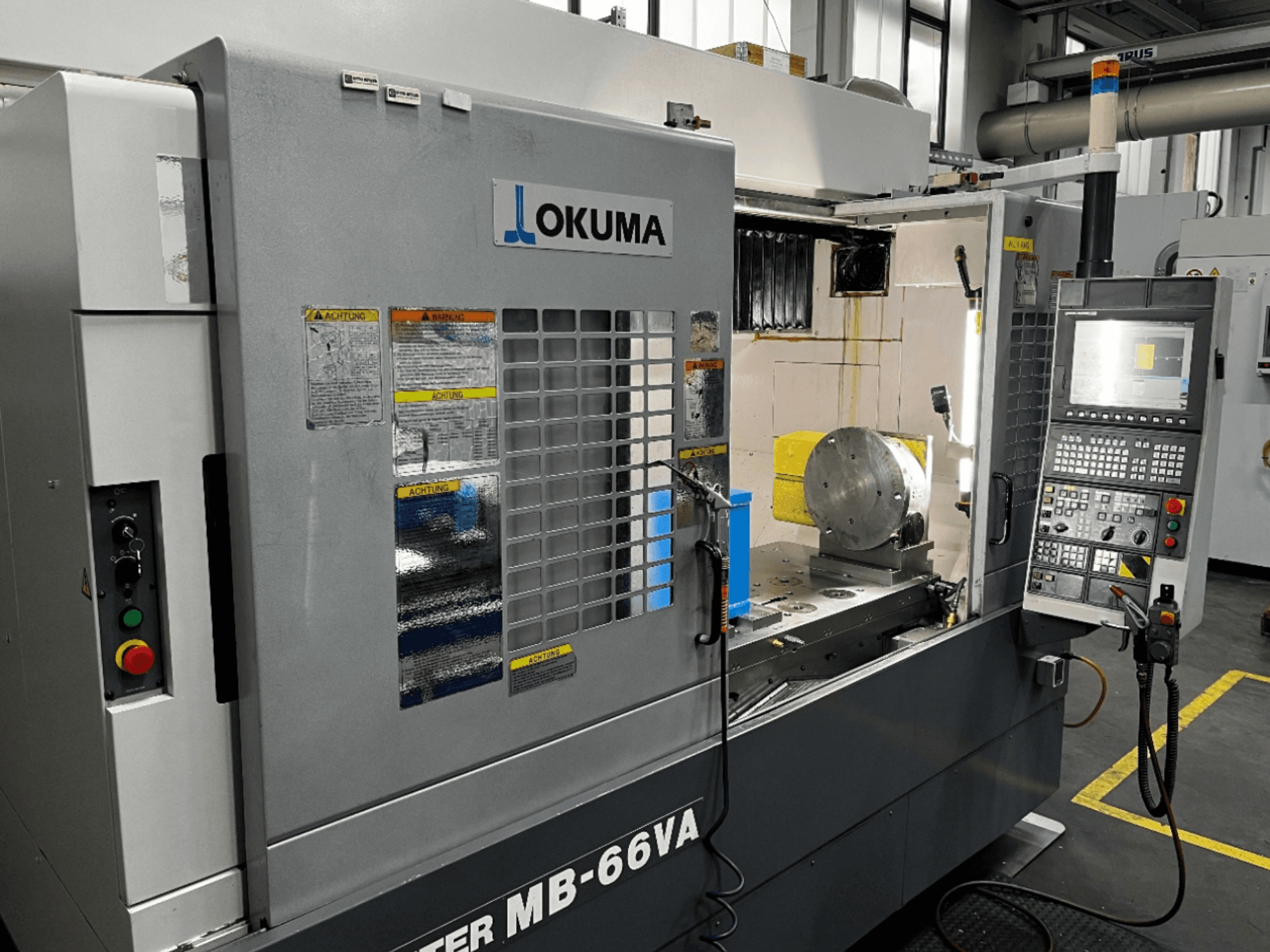 Okuma MB66-VA Vertical Machining Centre etukuva, jossa näkyy ohjauspaneeli, työkappale ja koneen sisätilat sekä turvatarrat.