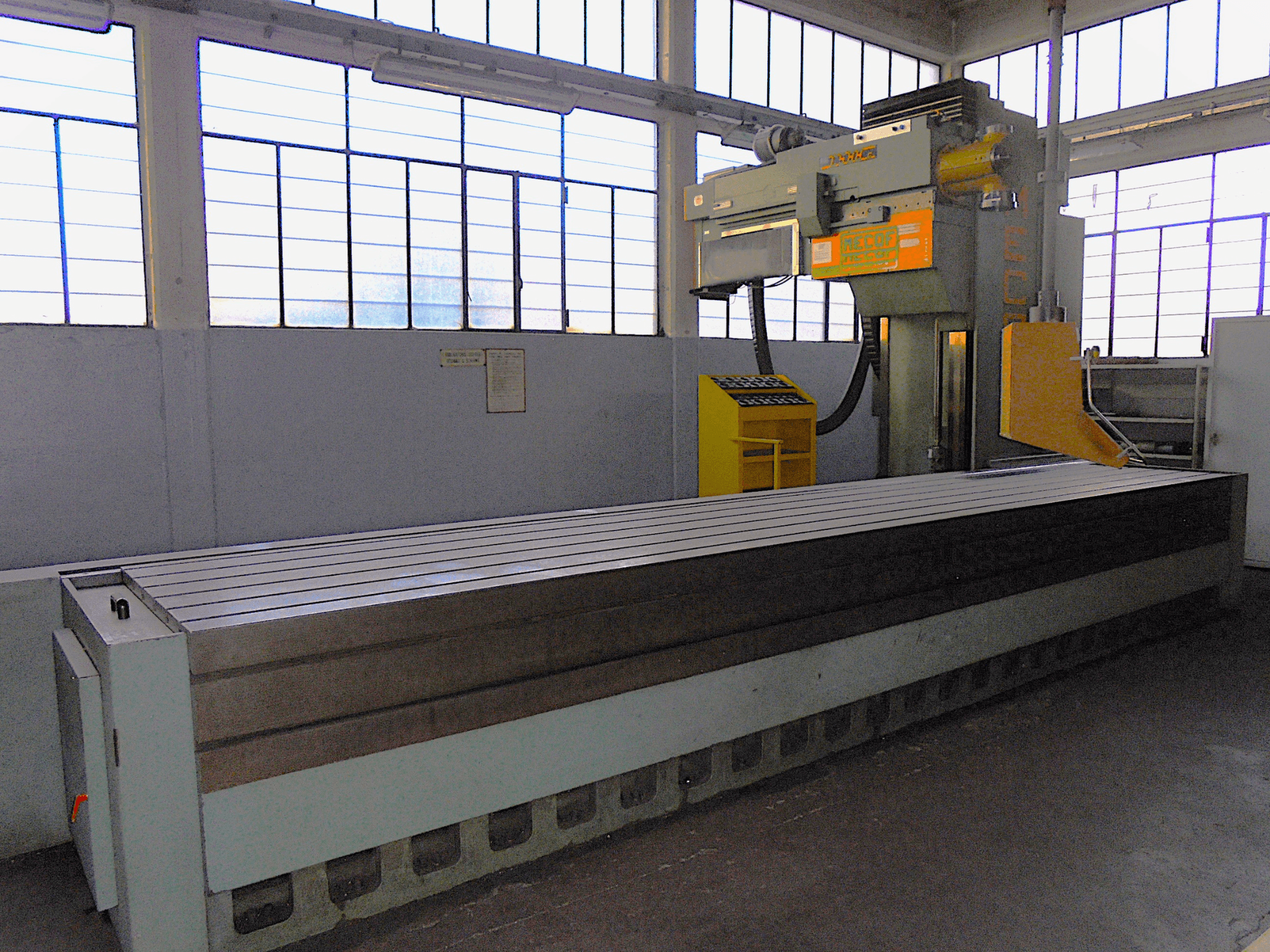 Merkki tuntematon, pystysuora jyrsinkone tehtaalla, sivukuva, jossa on suuri pöytä, jossa on uritettu pinta ja ohjauspaneeli.