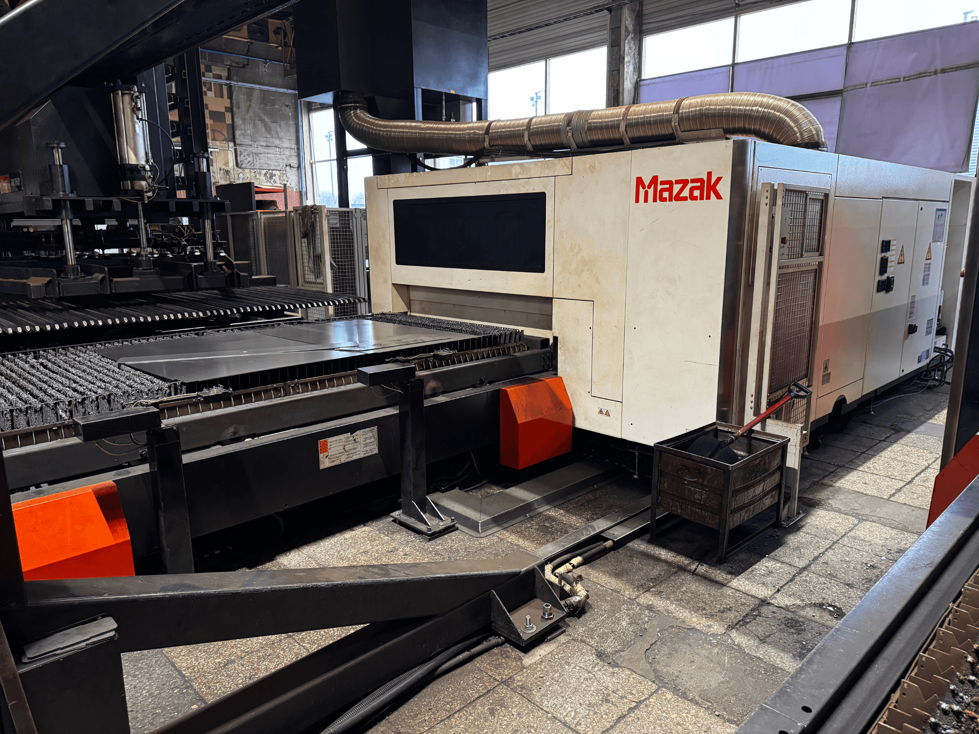 Mazak-laserleikkauskone työpajassa, sivukuva, jossa näkyy ohjauspaneeli, kuljetin ja keräysastia.