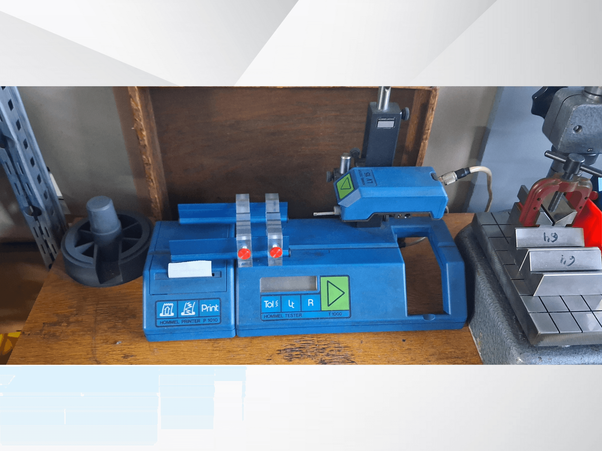 Sininen Hommel T 1000 -tulostin ja T 1 010 -testeri, jossa on punaiset painikkeet, edestä katsottuna metallipalikoilla varustetulla työpöydällä.