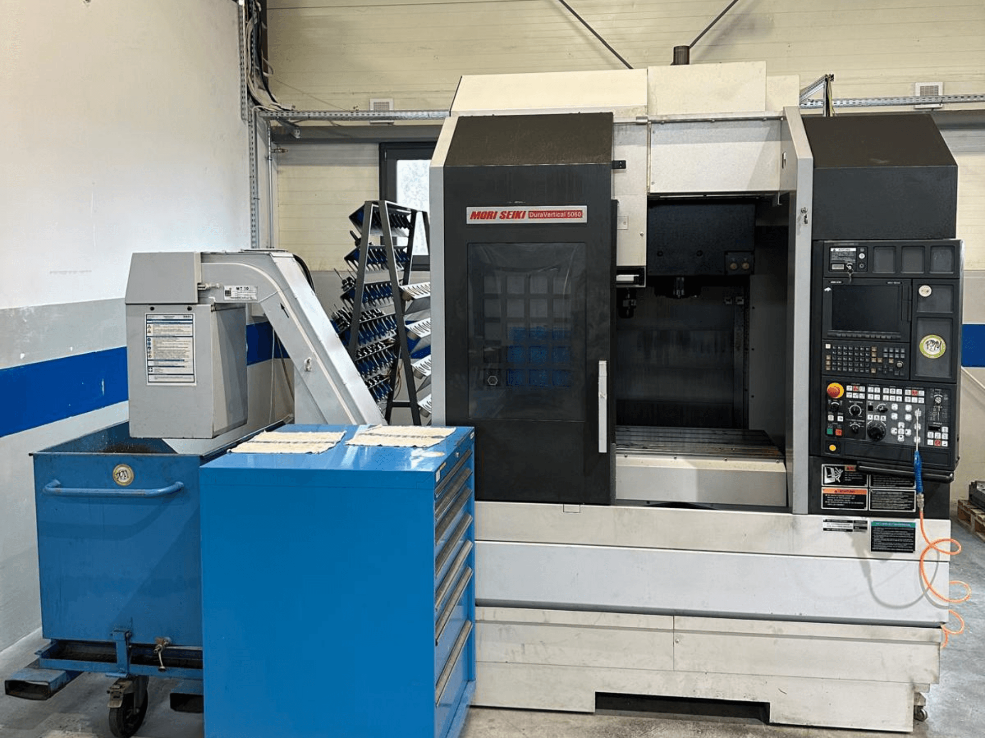MORI SEIKI DURA VERTICAL 5060, pystykaraisen työstökeskuksen etunäkymä, jossa näkyy ohjauspaneeli ja sen vieressä oleva työkaluvarasto.