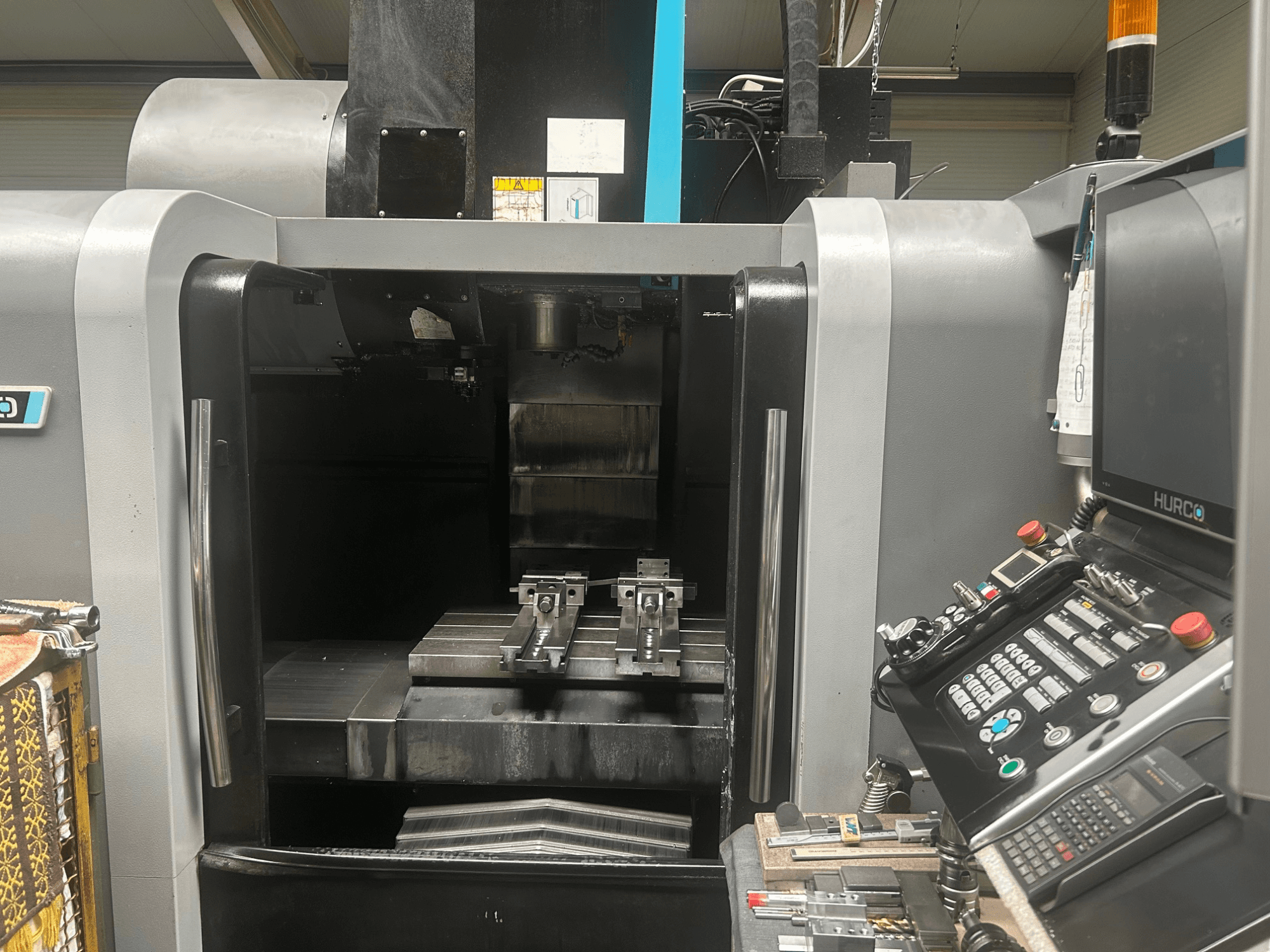 Hurco CNC-työstökeskuksen etukuva, jossa näkyy työkalunpidin, metallinen työkappale ja ohjauspaneeli, jossa on painikkeet ja näyttö.