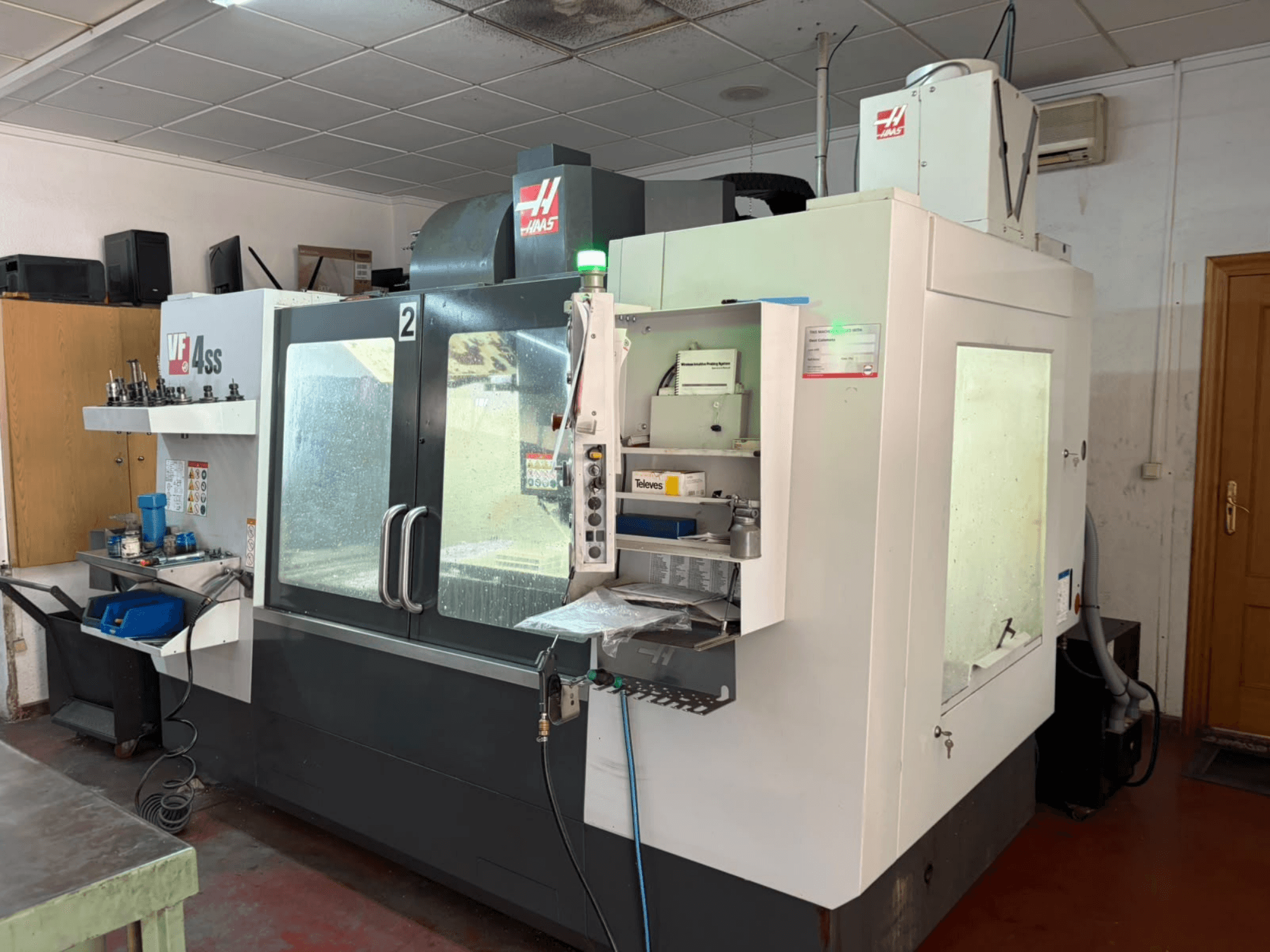HAAS VF 4SS -pystykoneistuskeskus työpajassa, etunäkymä, jossa näkyy ohjauspaneeli sekä työalue työkaluineen ja materiaaleineen.