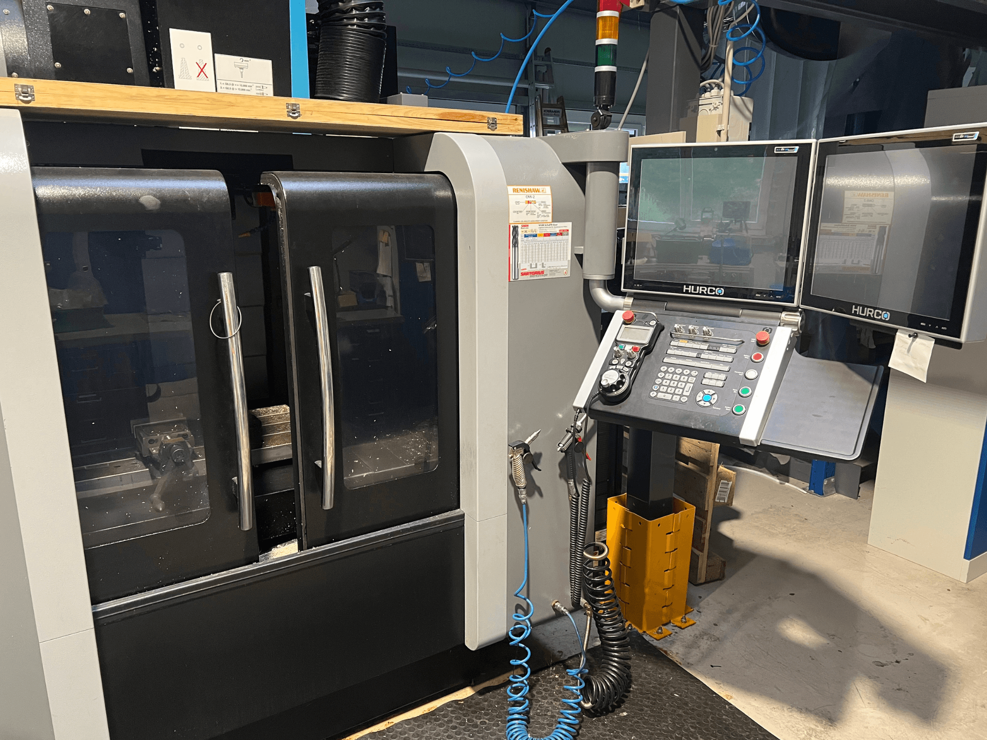 Hurco CNC-koneen etukuva, jossa on kaksi näyttöä ja ohjauspaneeli. Sisällä on läpinäkyvät ovet ja näkyvä työstöalue.