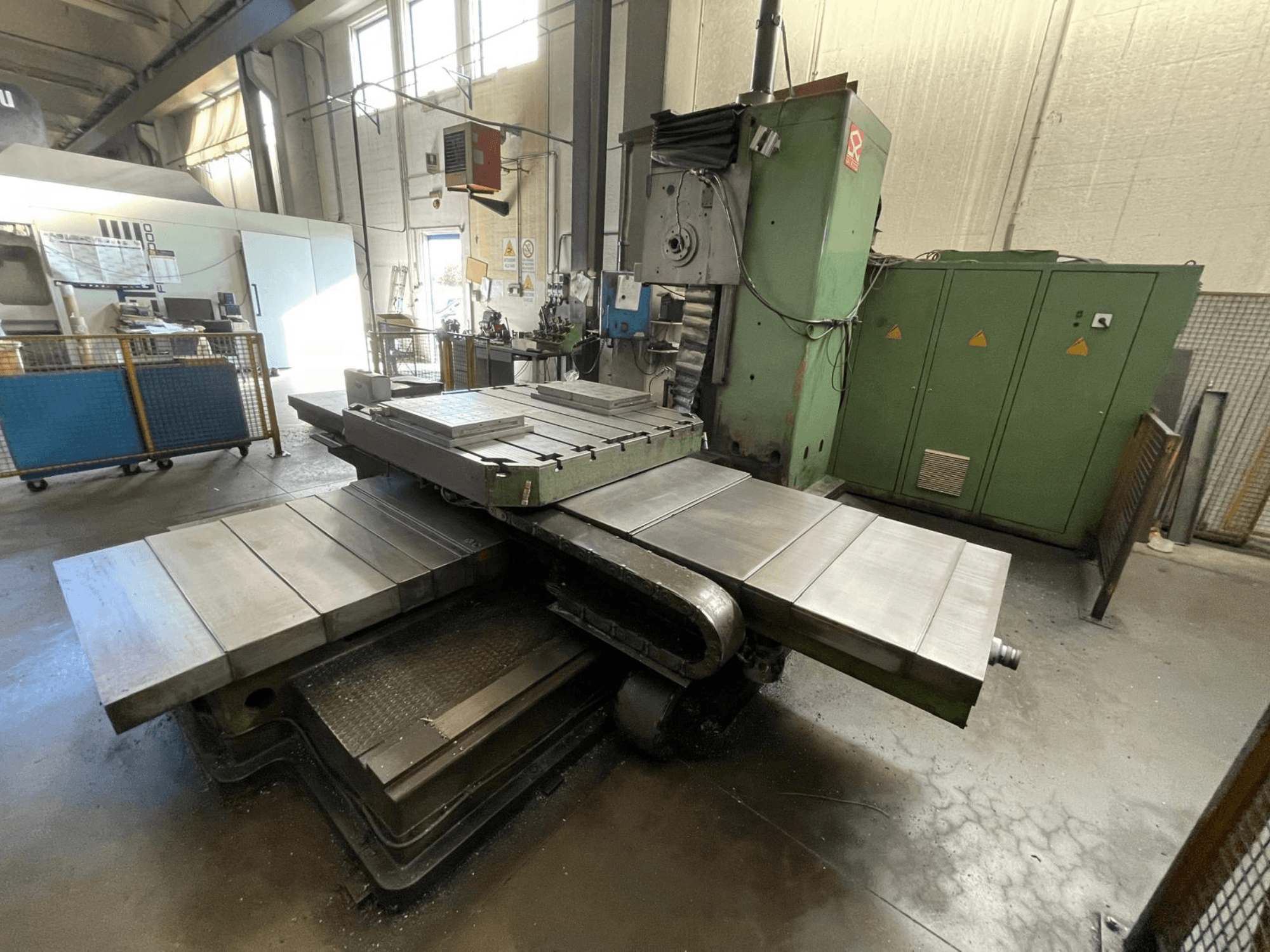 Suuri teollinen jyrsinkone, jossa on vihreä runko, sivukuva, jossa on useita tasaisia pintoja ja varastokaapit taustalla.