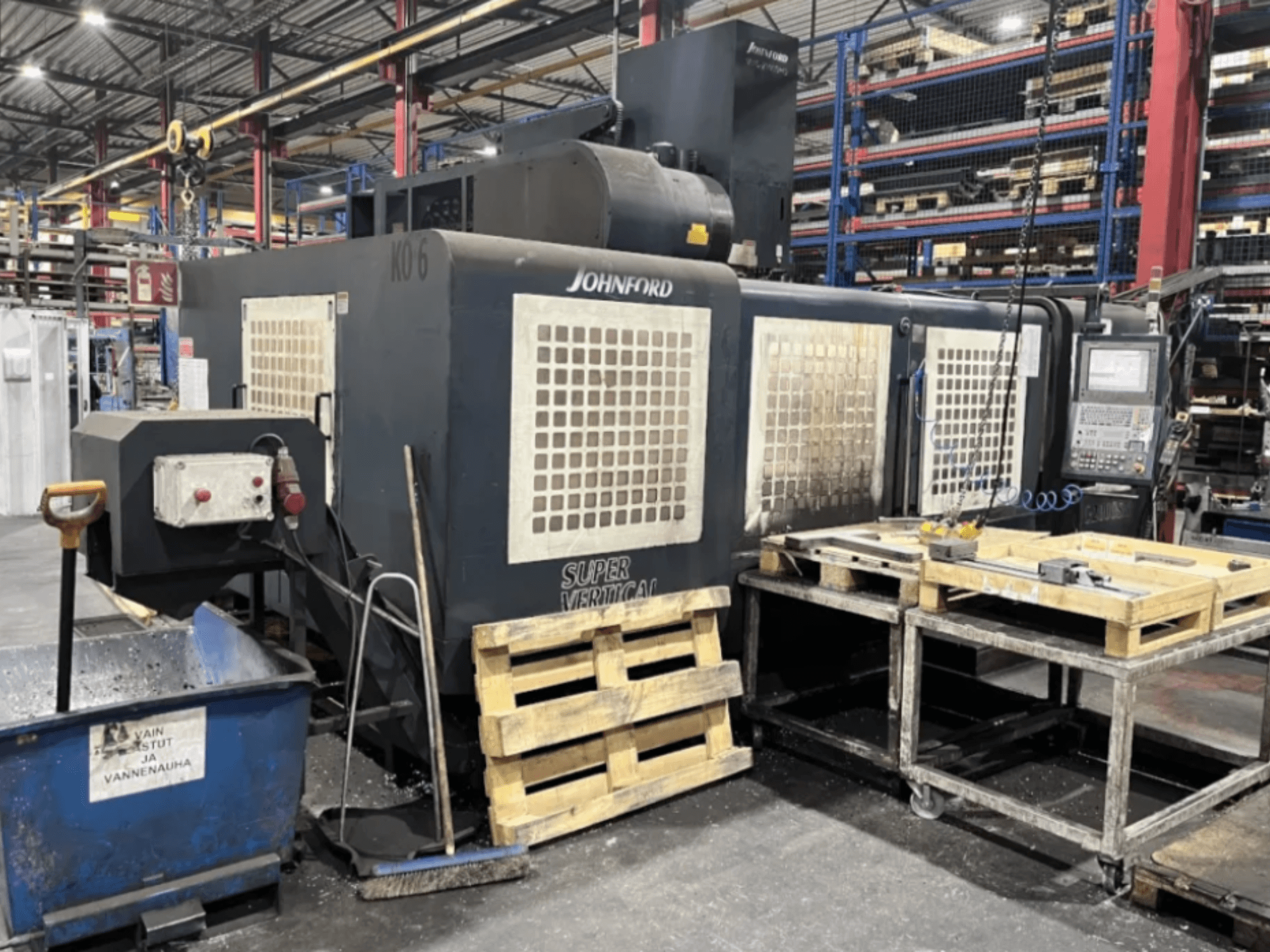 Johnford Super Vertical -jyrsinkone tehtaalla, sivukuva, jossa on ohjauspaneeli, kuormalavoja ja puhdistuslaitteita lähellä.