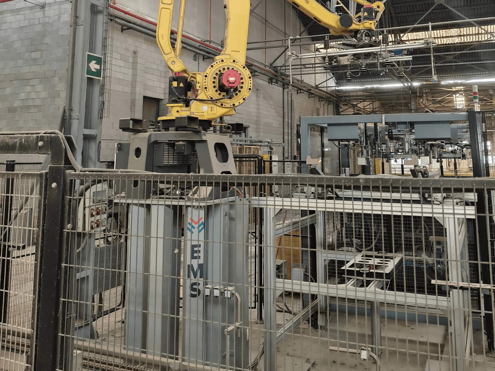 Etunäkymä FANUC M-410iC/315 -robottivarresta, joka on sijoitettu EMS-työkalun yläpuolelle; taustalla näkyy teollisuuslaitteita ja suojakaiteita.