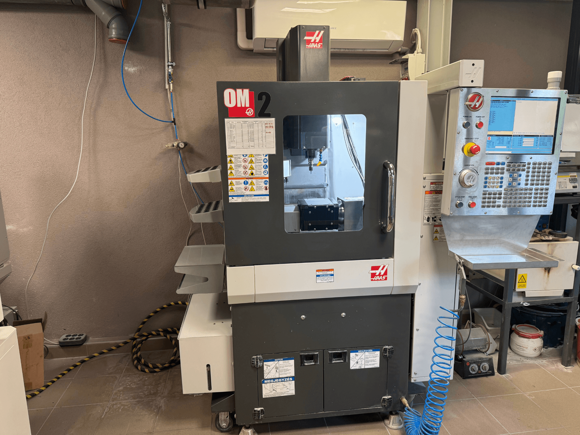 Haas OM-2 CNC-kone, etukuva, jossa on ohjauspaneeli, kara ja turvatarrat, asetettu työpajaympäristöön.