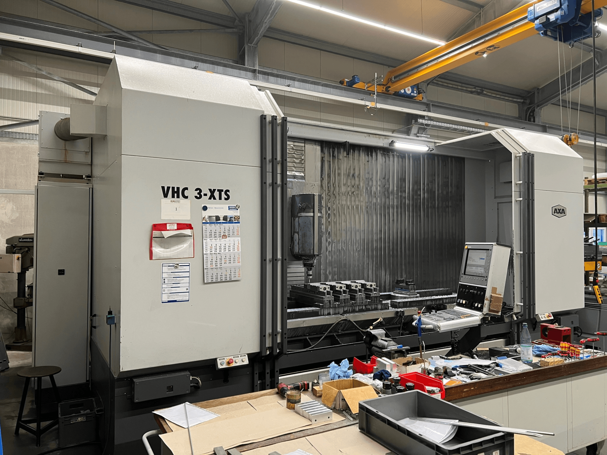 AXA VHC 3-XTS CNC-kone sivukuvassa, ohjauspaneeli ja työalue täynnä työkaluja ja komponentteja.