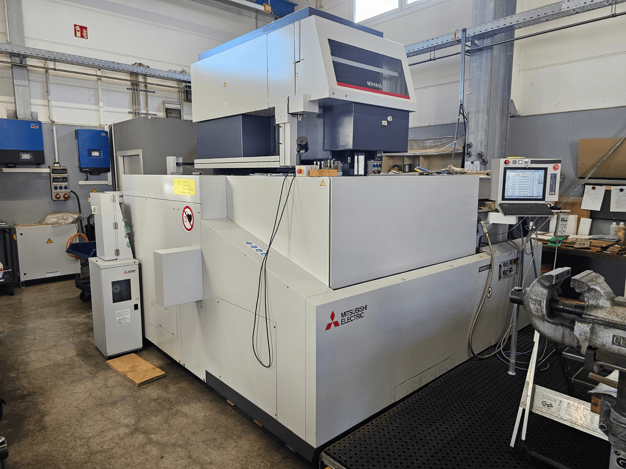 Mitsubishi MV4800 Wire EDM -kone työpajassa, sivukuva, jossa näkyy ohjauspaneeli, turvatarrat ja ympäröivät laitteet.