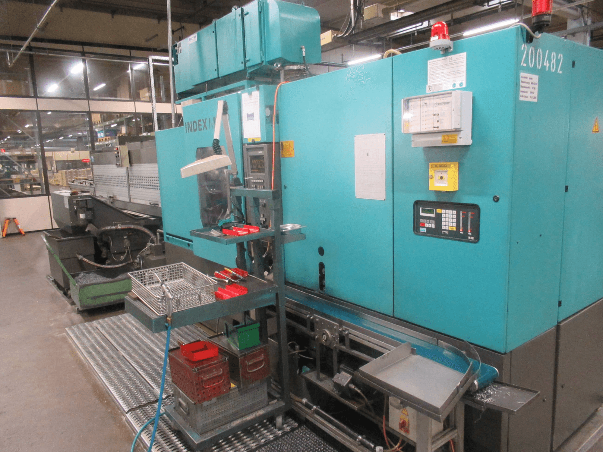Turkoosi INDEX-kone tehtaalla, sivukuva; ohjauspaneeli, työkalujen säilytys ja kuljetin. Näkyvät säiliöt, letkut ja hälytyslaitteet.
