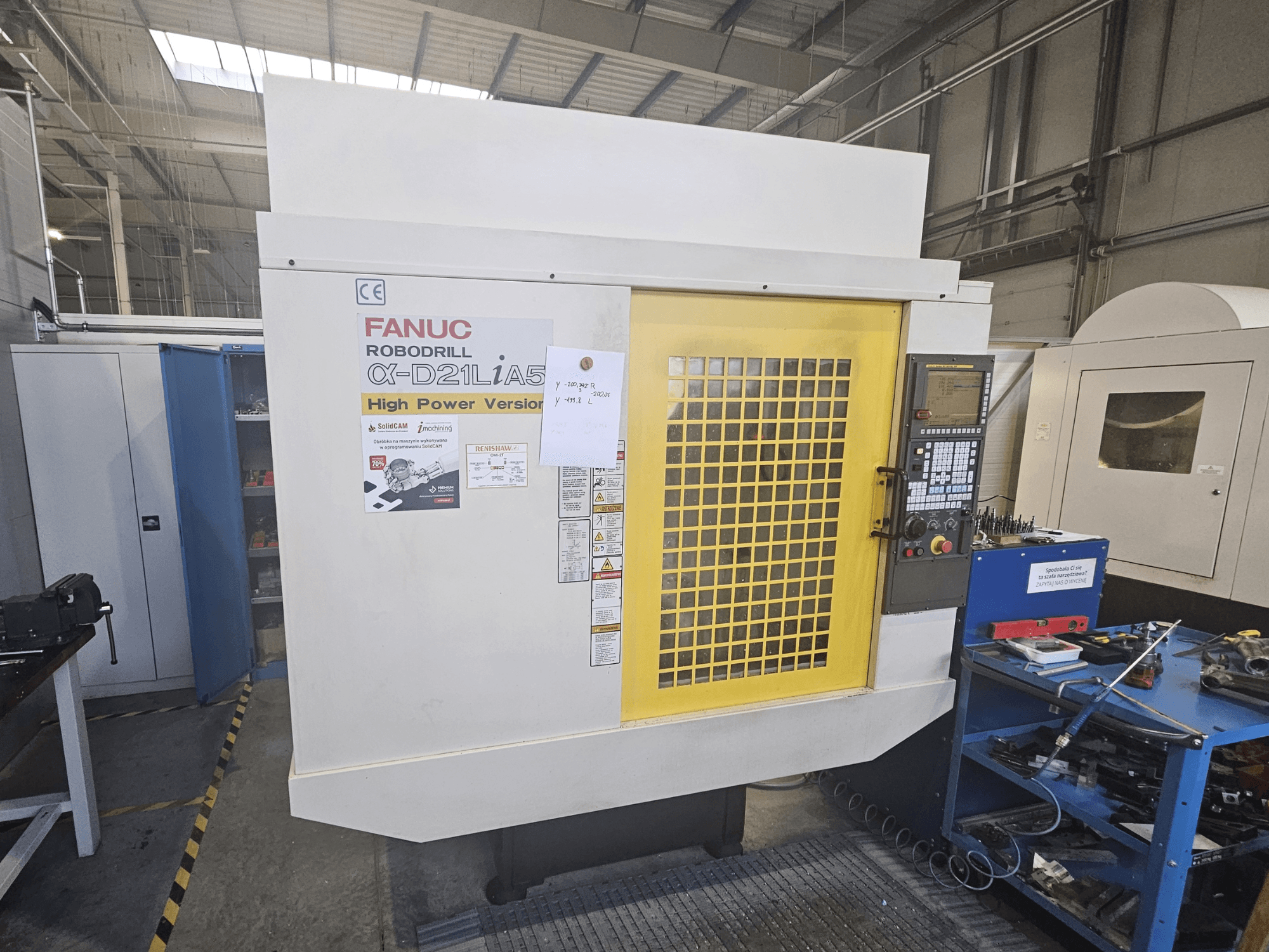FANUC Robodrill α-D21LiA5:n etukuva, jossa on keltainen turvaluukku, ohjauspaneeli ja työtilan työkalut lähellä.