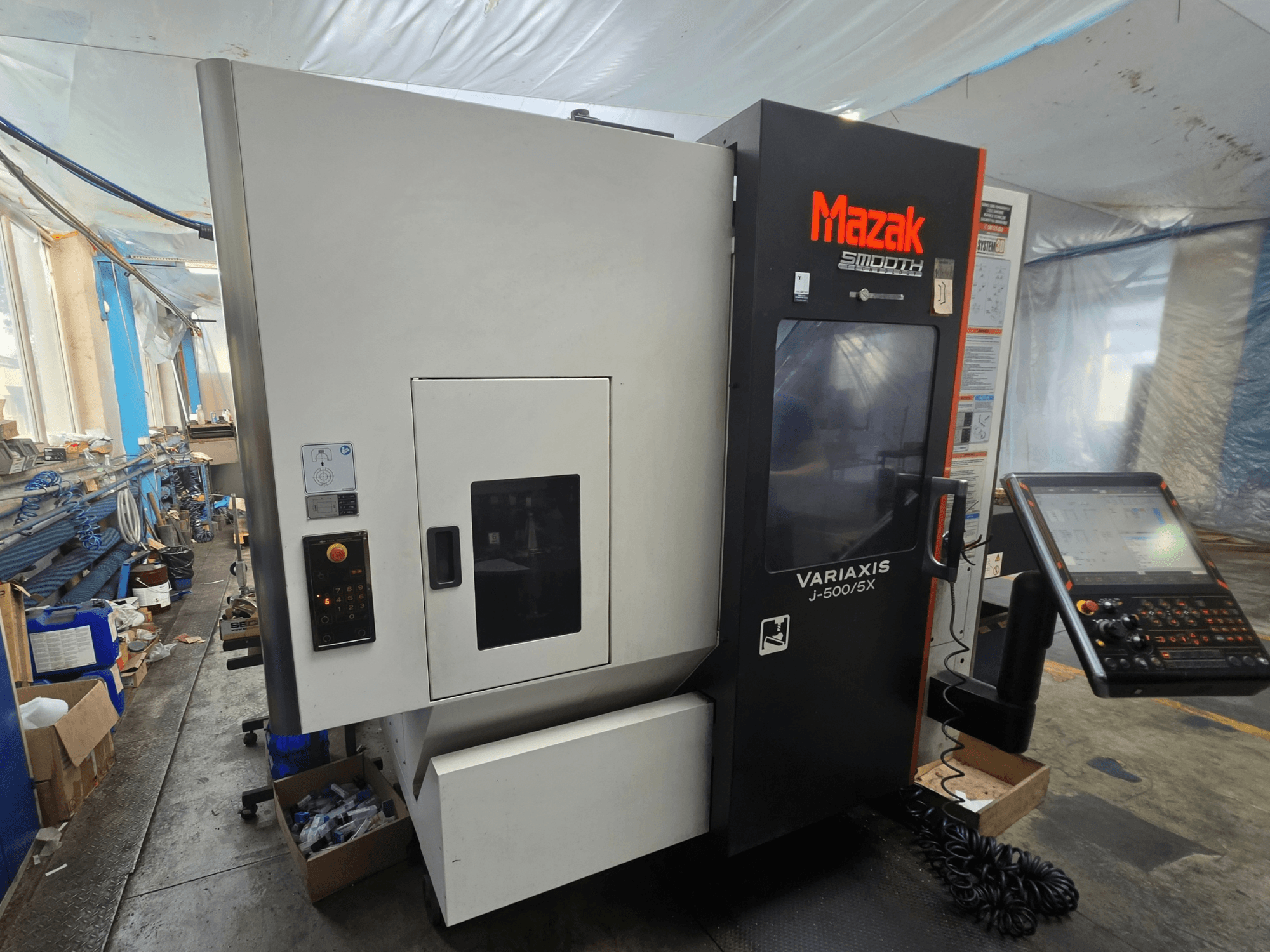 Mazak VARIAXIS J-500/5X -pystykoneistuskeskus, sivukuva, jossa näkyvät hallintalaitteet ja näyttö, sijoitettuna työpajaympäristöön.
