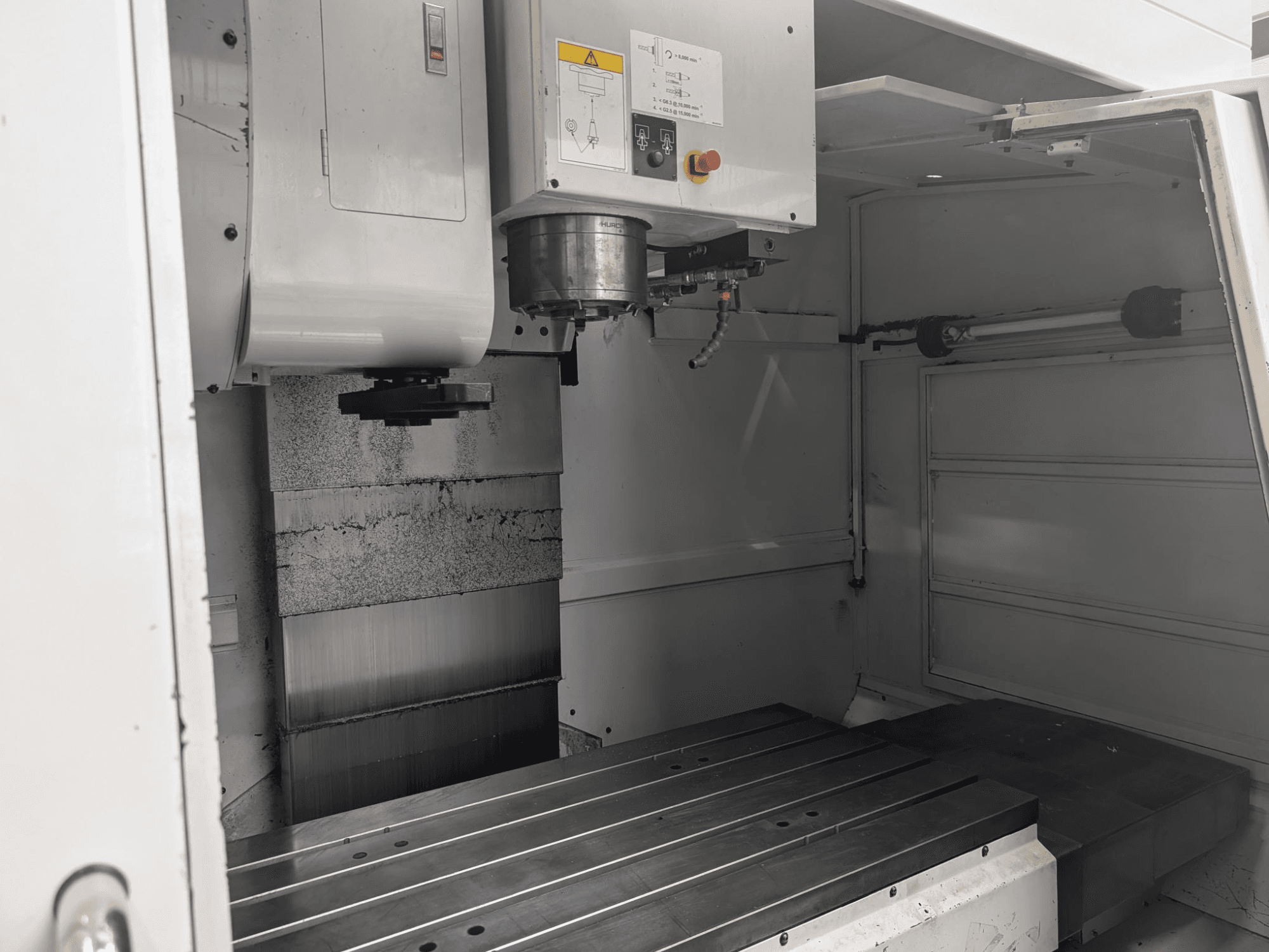 Sisäkuva CNC-jyrsinkoneesta, jossa on suuri metallinen työpöytä ja karamoottori, jossa näkyy yksityiskohtaisia osia ja ohjauspaneeli.