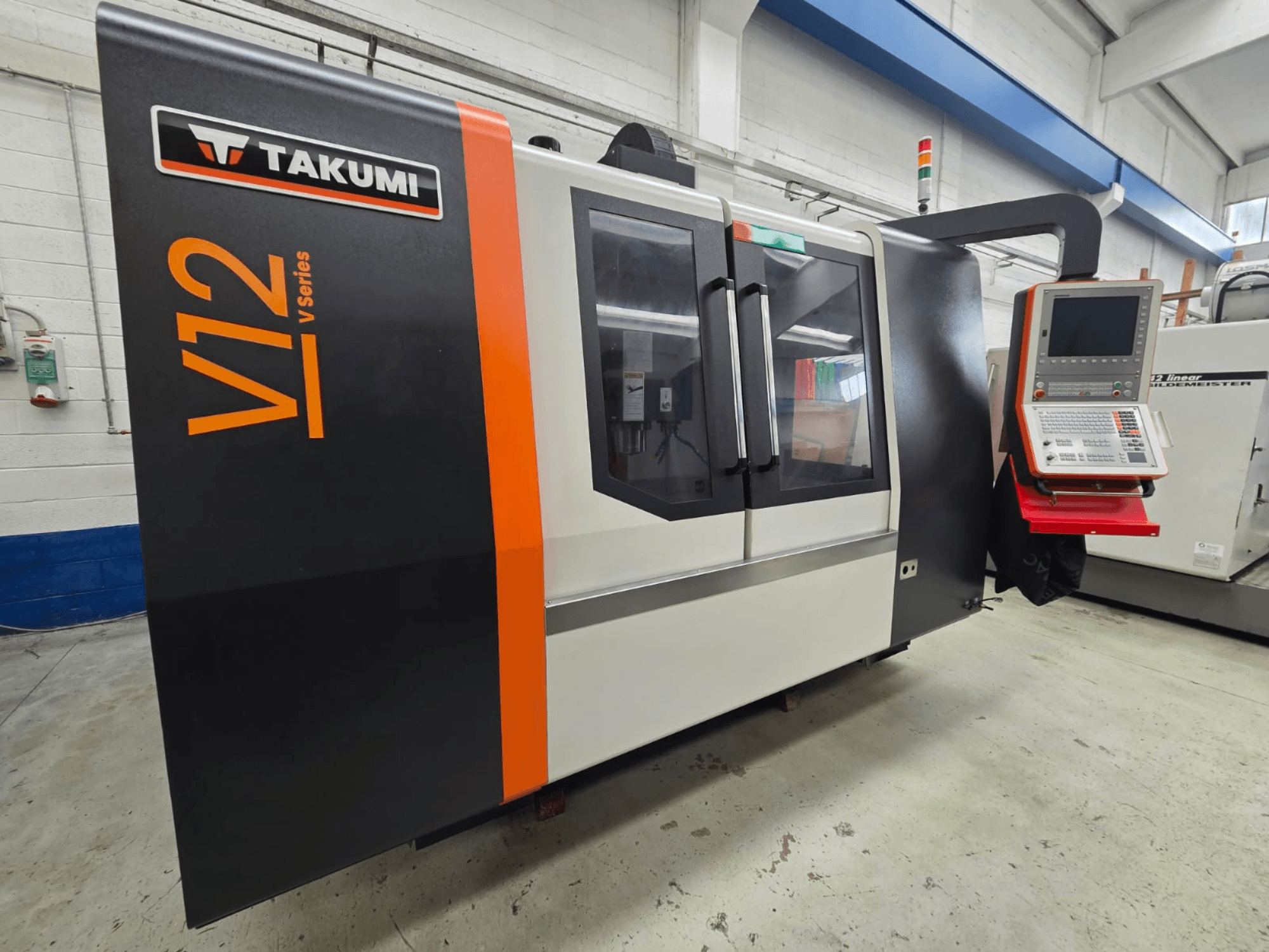 Takumi V12 V-sarjan CNC-jyrsin, etukuva, jossa on ohjauspaneeli ja suuret lasi-ikkunat.