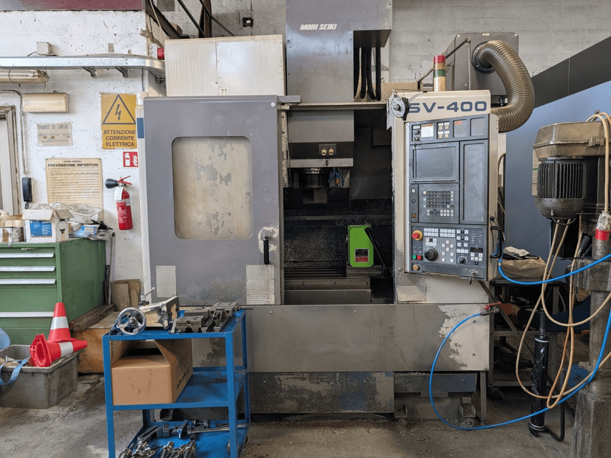 MORI SEIKI SV-400 -pystytyöstökeskus työpajassa, sivukuva, jossa näkyy ohjauspaneeli ja työstökoneet lähellä.