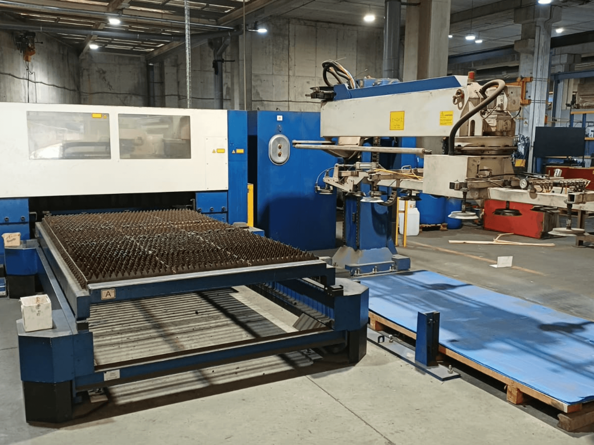 TRUMPF TRUMATIC L 3030 CO₂-laserleikkauskone, sivukuva, jossa näkyy piikkipintaisella leikkuupöydällä varustettu kone ja robottivarsi.