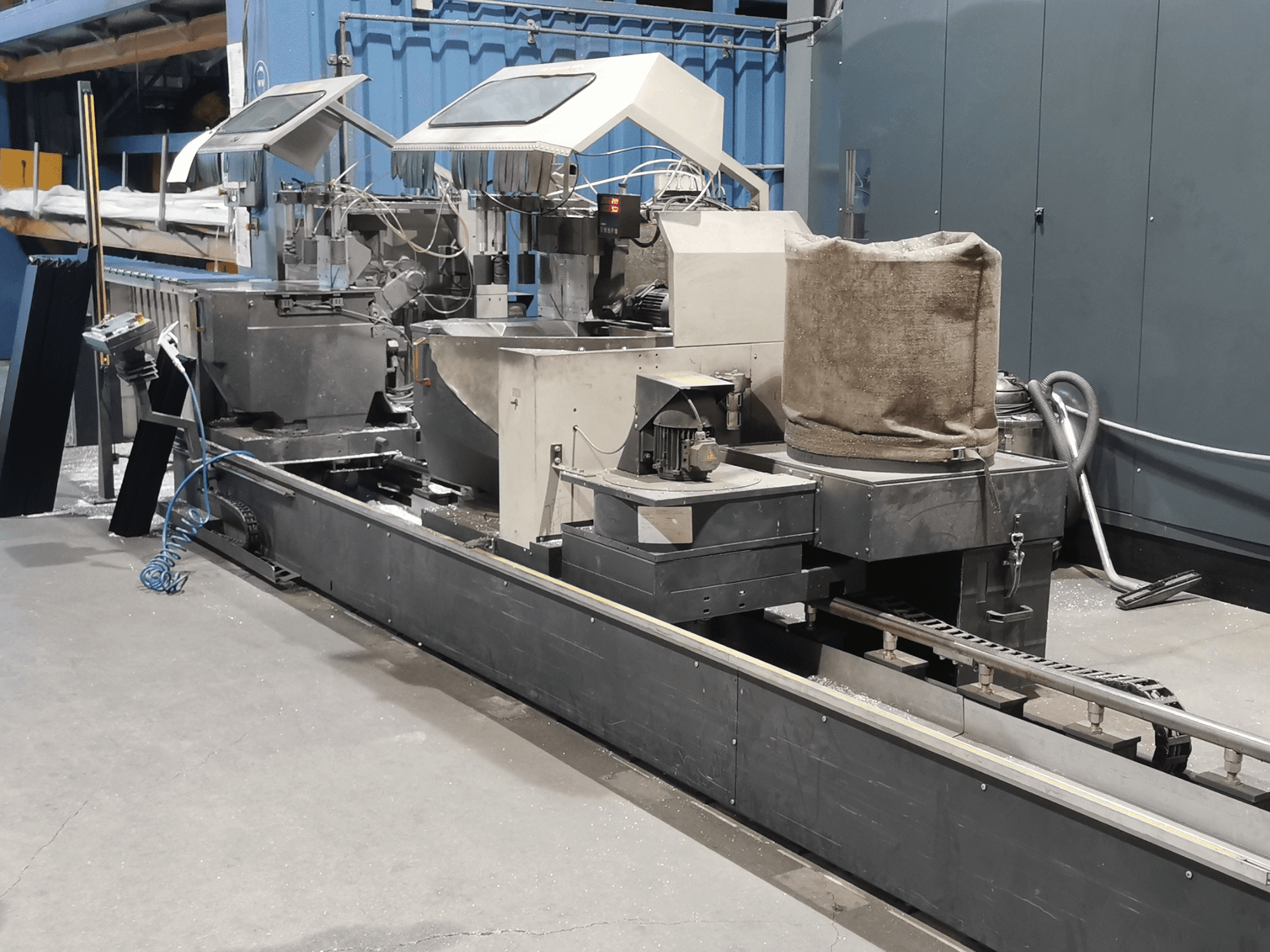 Teollinen käsittelykone sivulta katsottuna, jossa on suppiloita, kuljetinhihna ja ohjauspaneeli tehdasympäristössä.