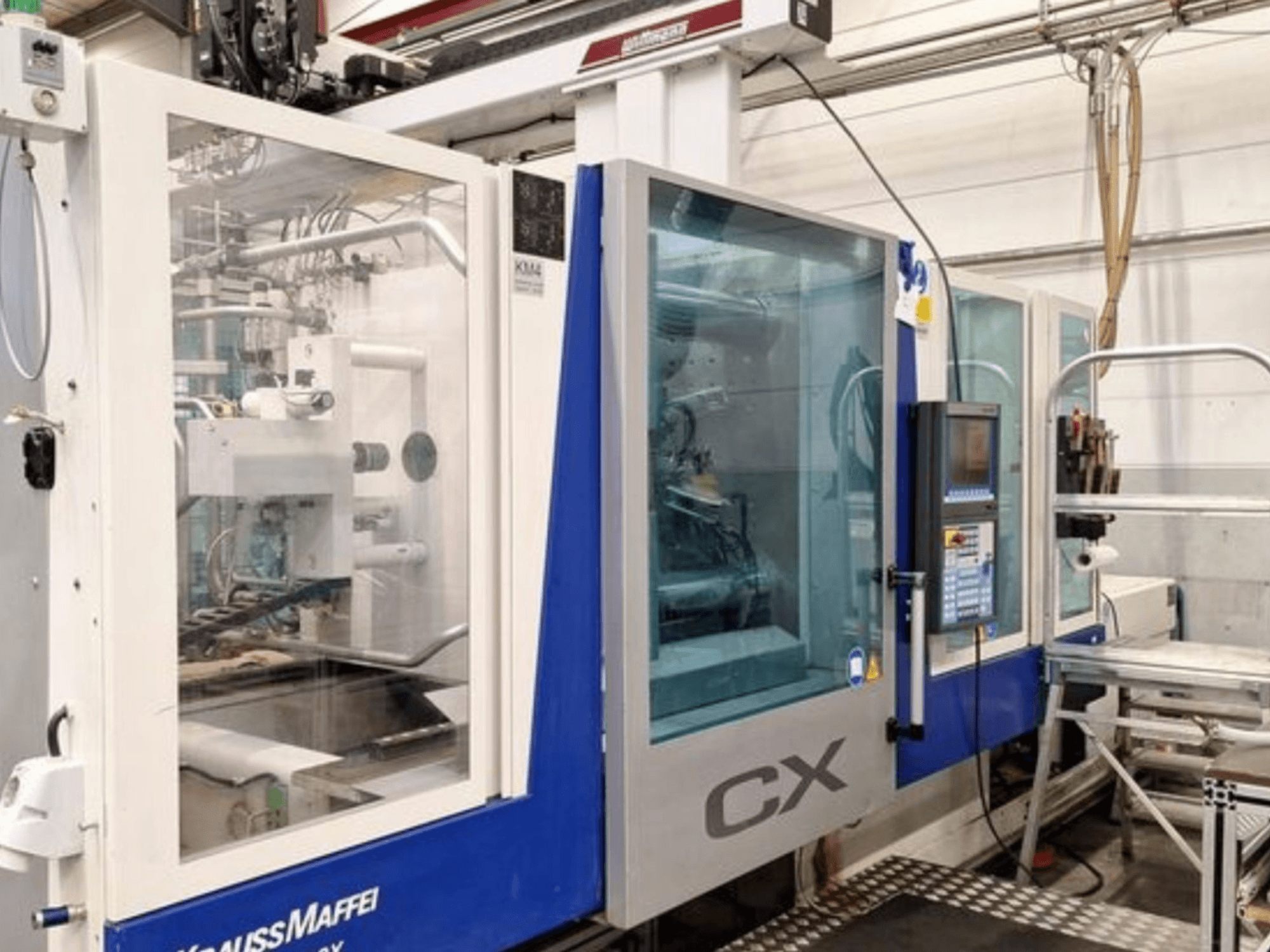 Krauss Maffei 300-1400 CX -hydraulinen ruiskuvalukone, sivukuva, jossa näkyvät ohjauspaneeli ja läpinäkyvä kotelo.