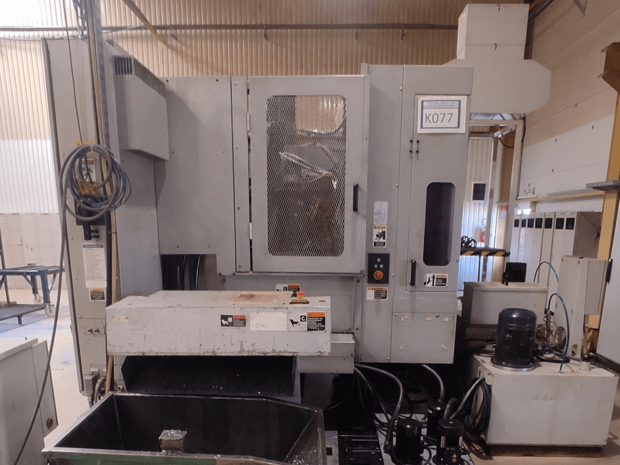 Harmaa teollinen CNC-kone, jossa on suuri työpöytä, sivukuva, jossa on ohjauspaneeli ja jäähdytysnestesäiliö, malli K077.
