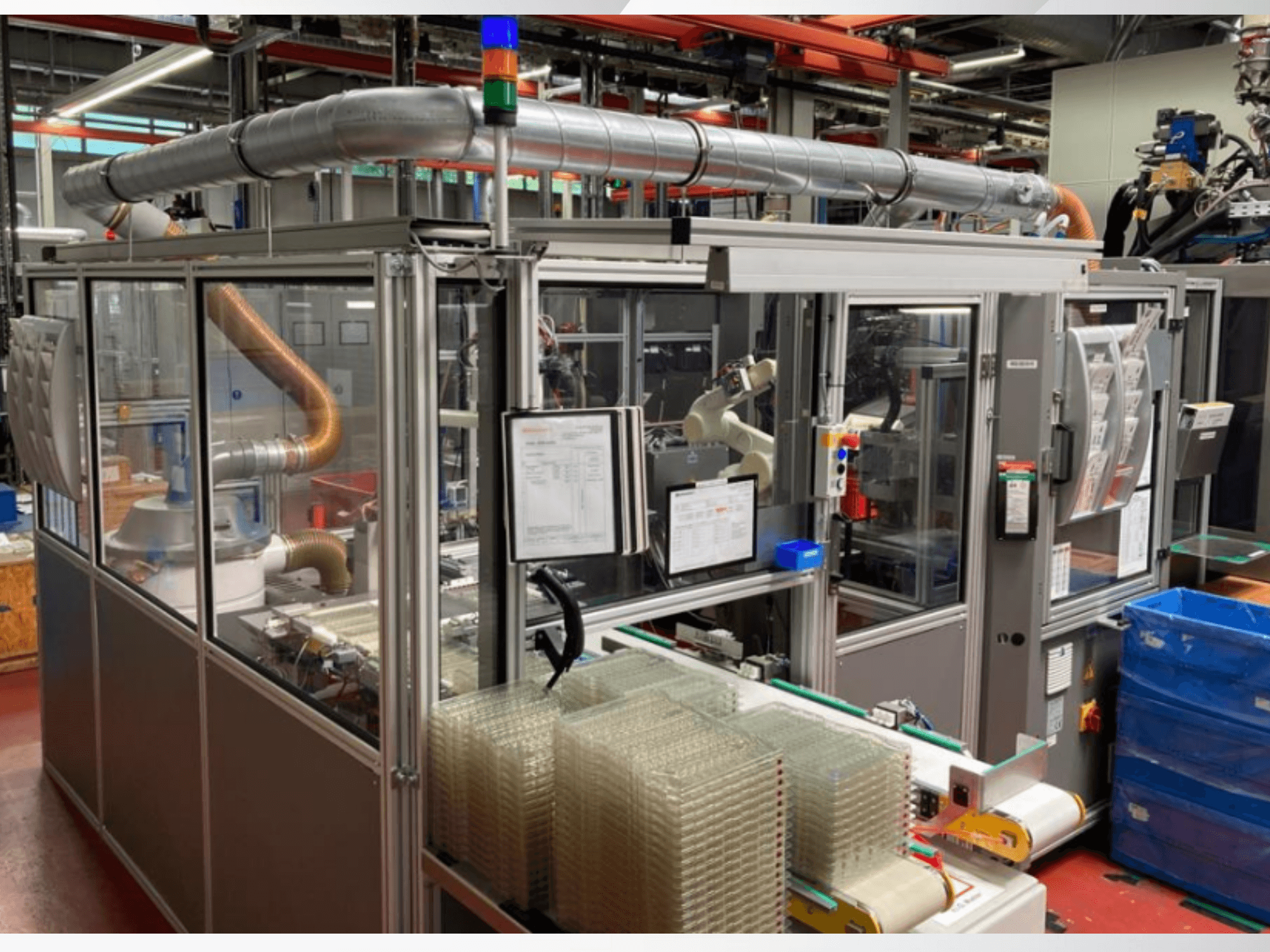 Etukuva automatisoidusta koneesta, jossa on metalliputkia, ohjauspaneeli ja pinottuja kirkkaita muovisäiliöitä tehtaan lattialla.