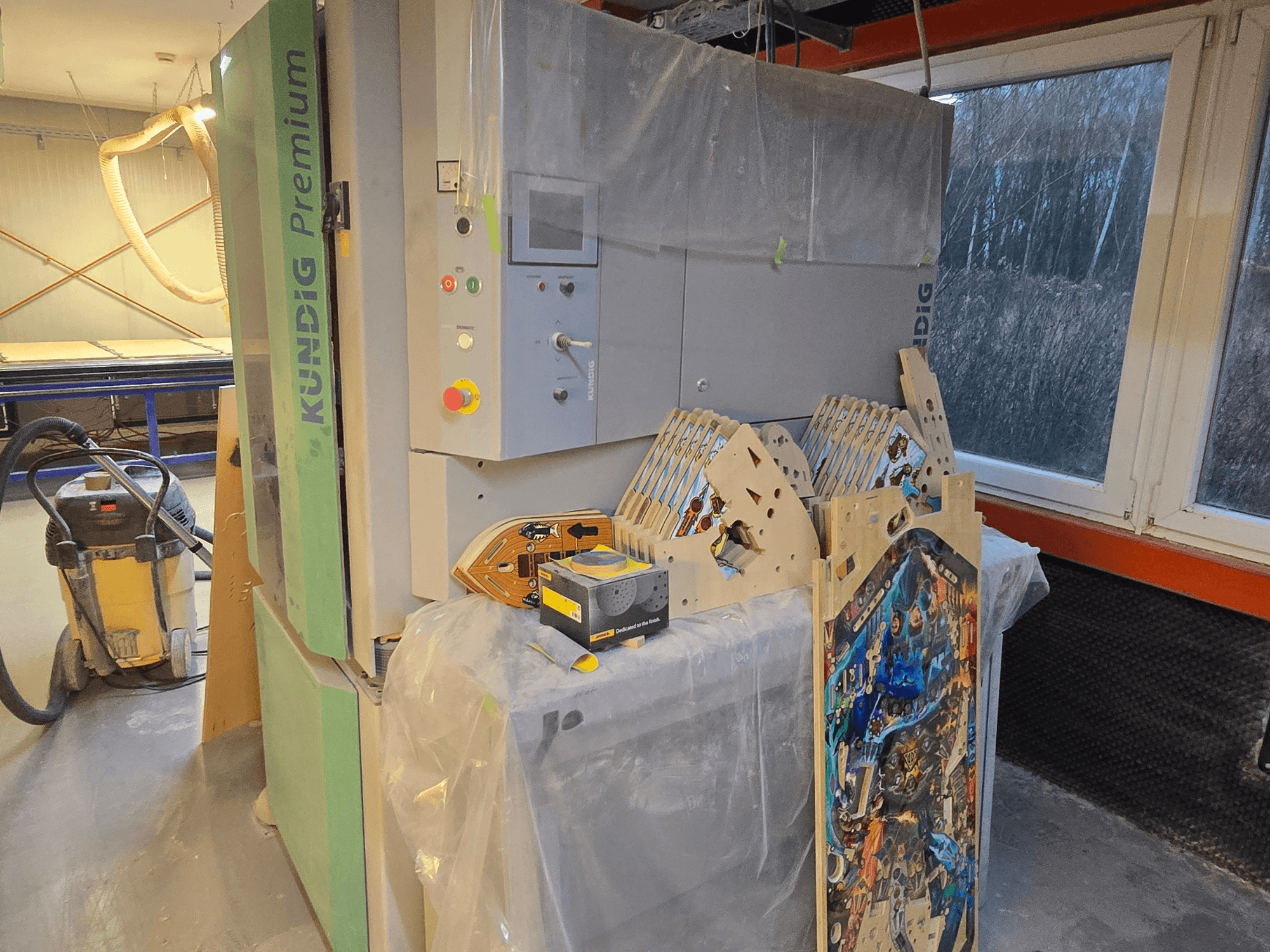 Kundig Premium-1 1100-C -hihnahiomakone työpajassa, ohjauspaneeli ja puukappaleet, sivukuva, taustalla pölyimuri.