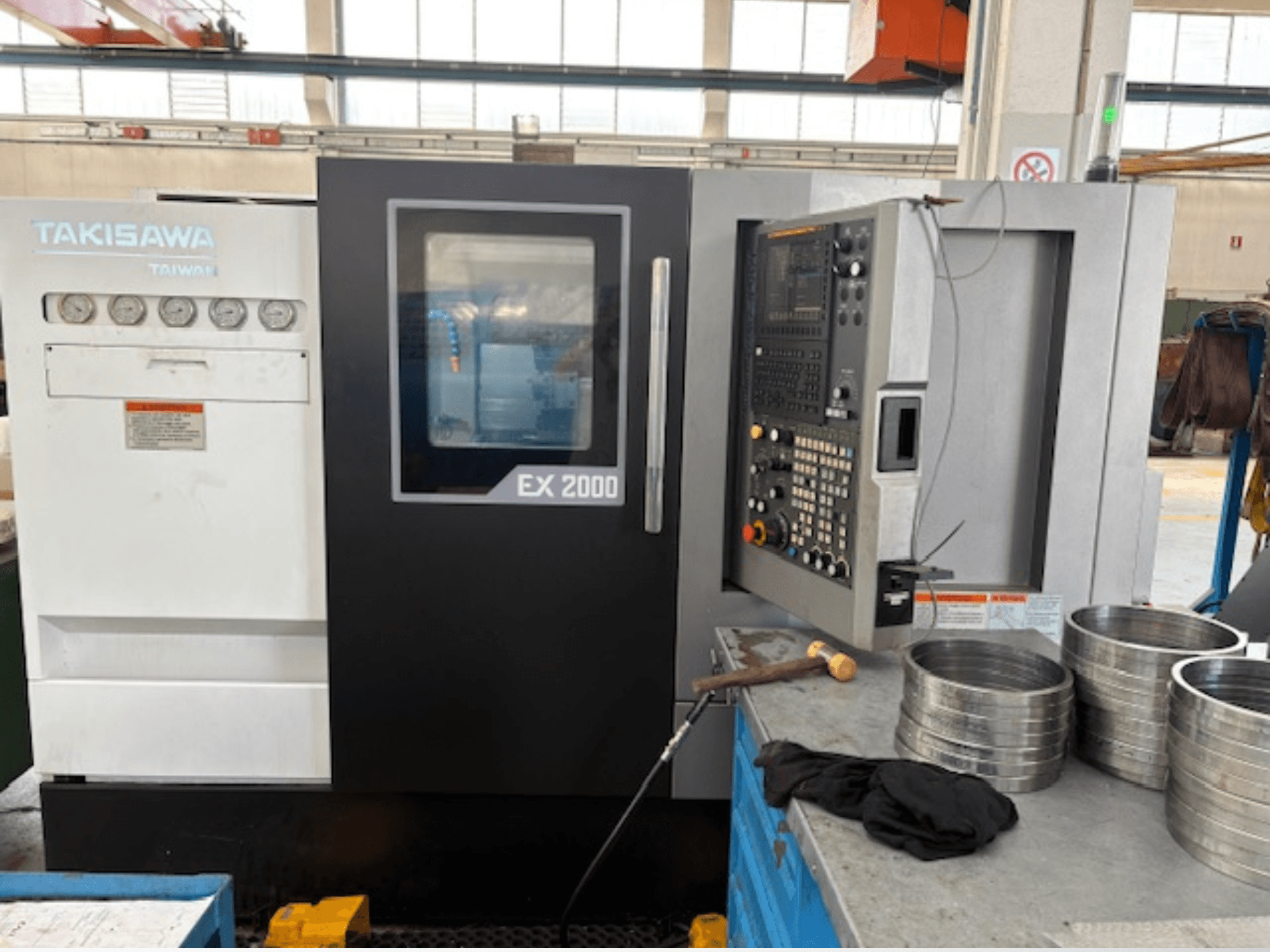 Takisawa EX 2000 CNC-kone, etukuva, jossa ohjauspaneeli, mittarit ja metalliosat työpöydällä.