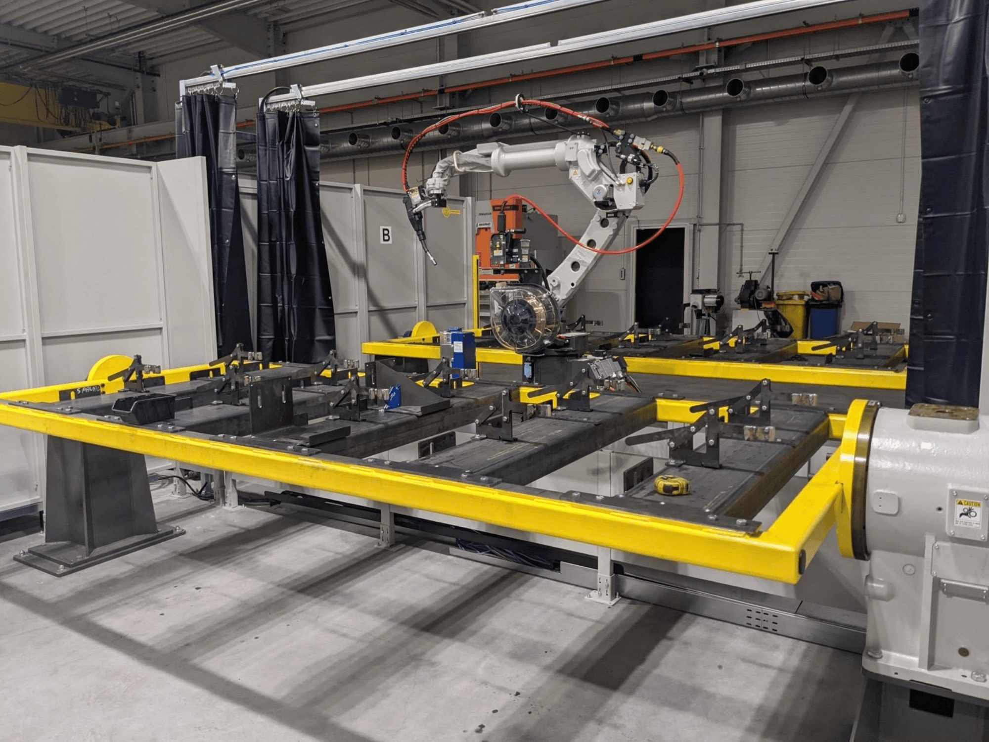 Etunäkymä PANASONIC TAWERS TM-2000WG3 -robottikäsivarresta, joka on sijoitettu keltaisen työpöydän yläpuolelle, jolla on kokoonpantavia metalliosia.