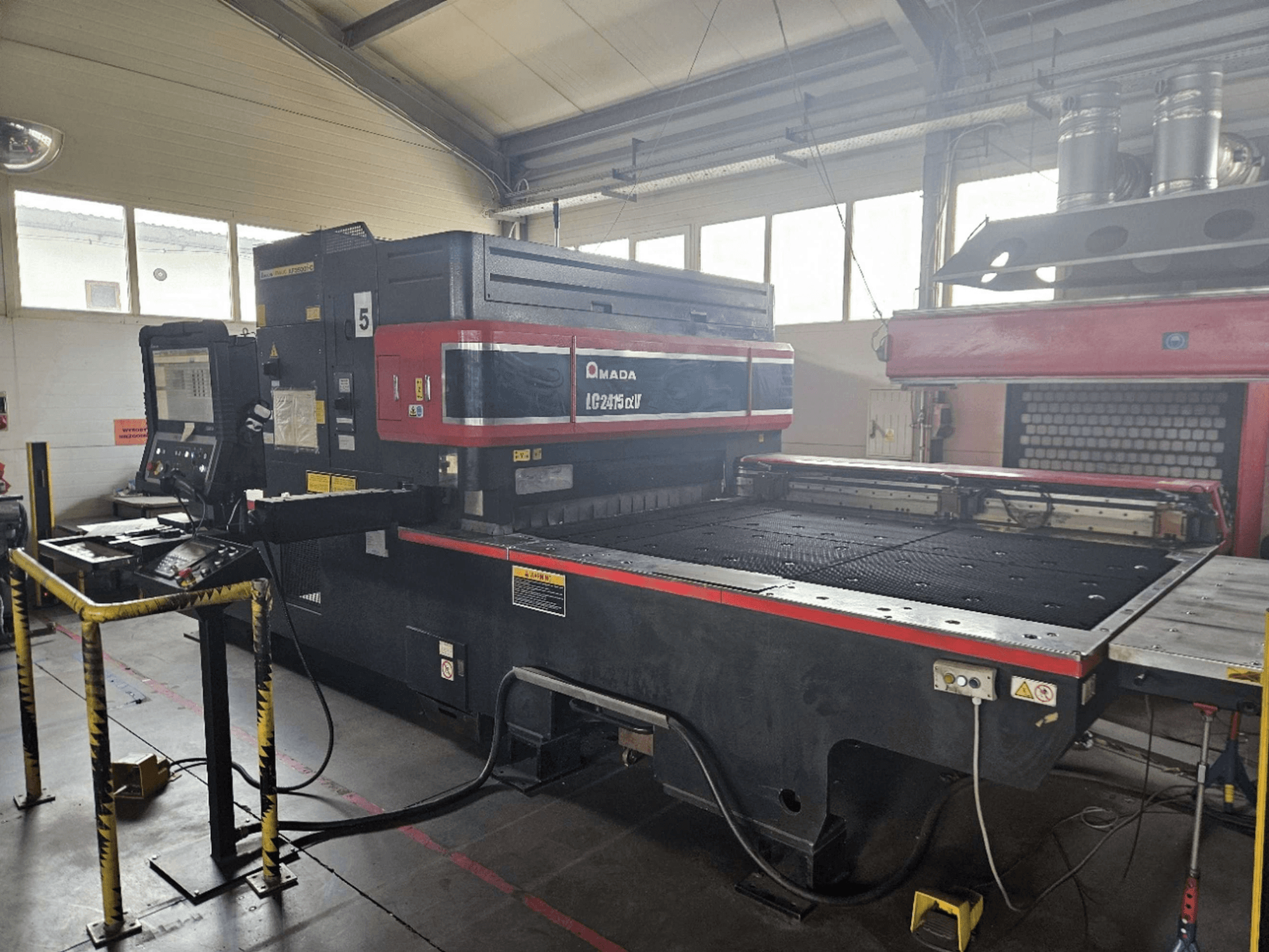 AMADA LC2415 Alpha 5 CO2-laserleikkauskone, etunäkymä, jossa näkyvät ohjauspaneeli ja leikkausalusta turva-aitoineen.