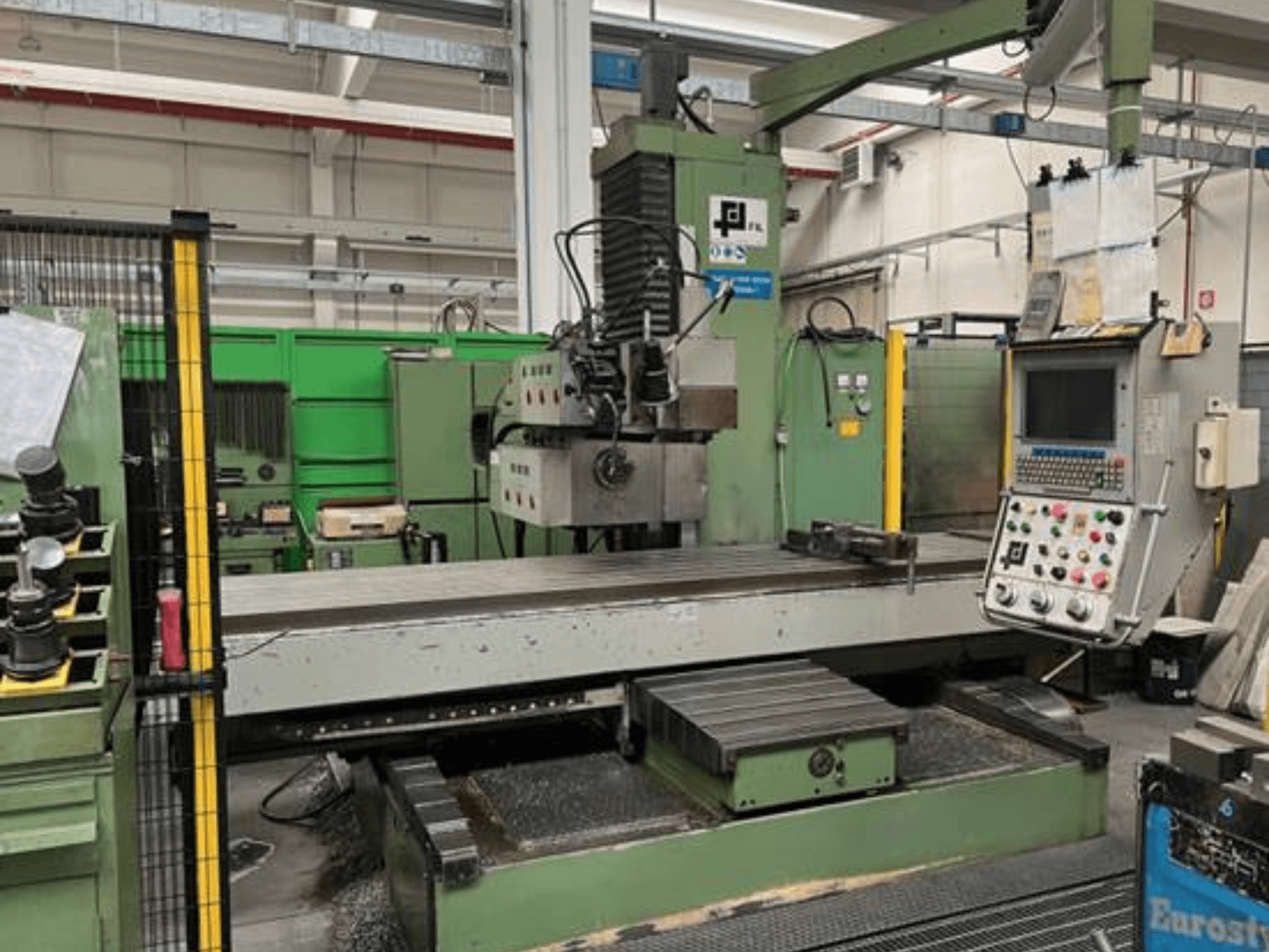 JFL:n teollisuusjyrsin, etukuva, jossa ohjauspaneeli, suuri tasainen pöytä ja metallilastuja lattialla.