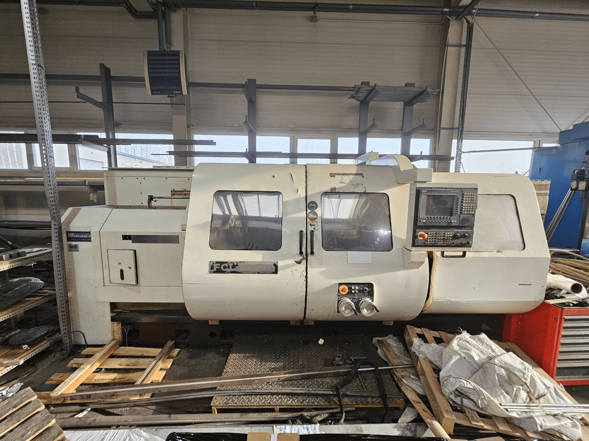 Wabeco CNC-sorvi, sivukuva, jossa ohjauspaneeli, ikkunat ja työskentelyalue; koneet puulavoilla työpajan ympäristössä.