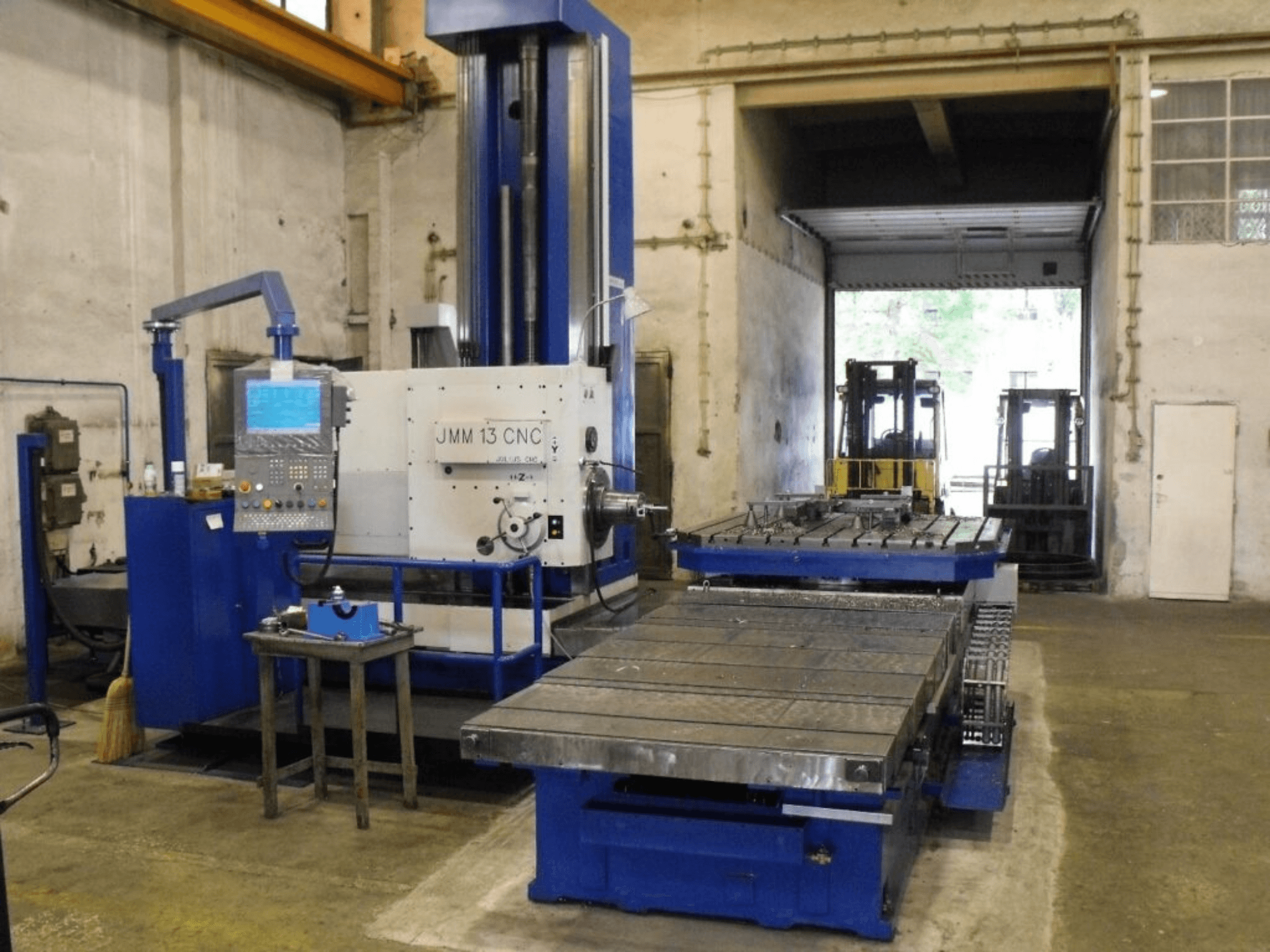 JMM 13 CNC-jyrsinkone työpajassa, etukuva, jossa on ohjauspaneeli, suuri työpöytä ja teollinen tausta.