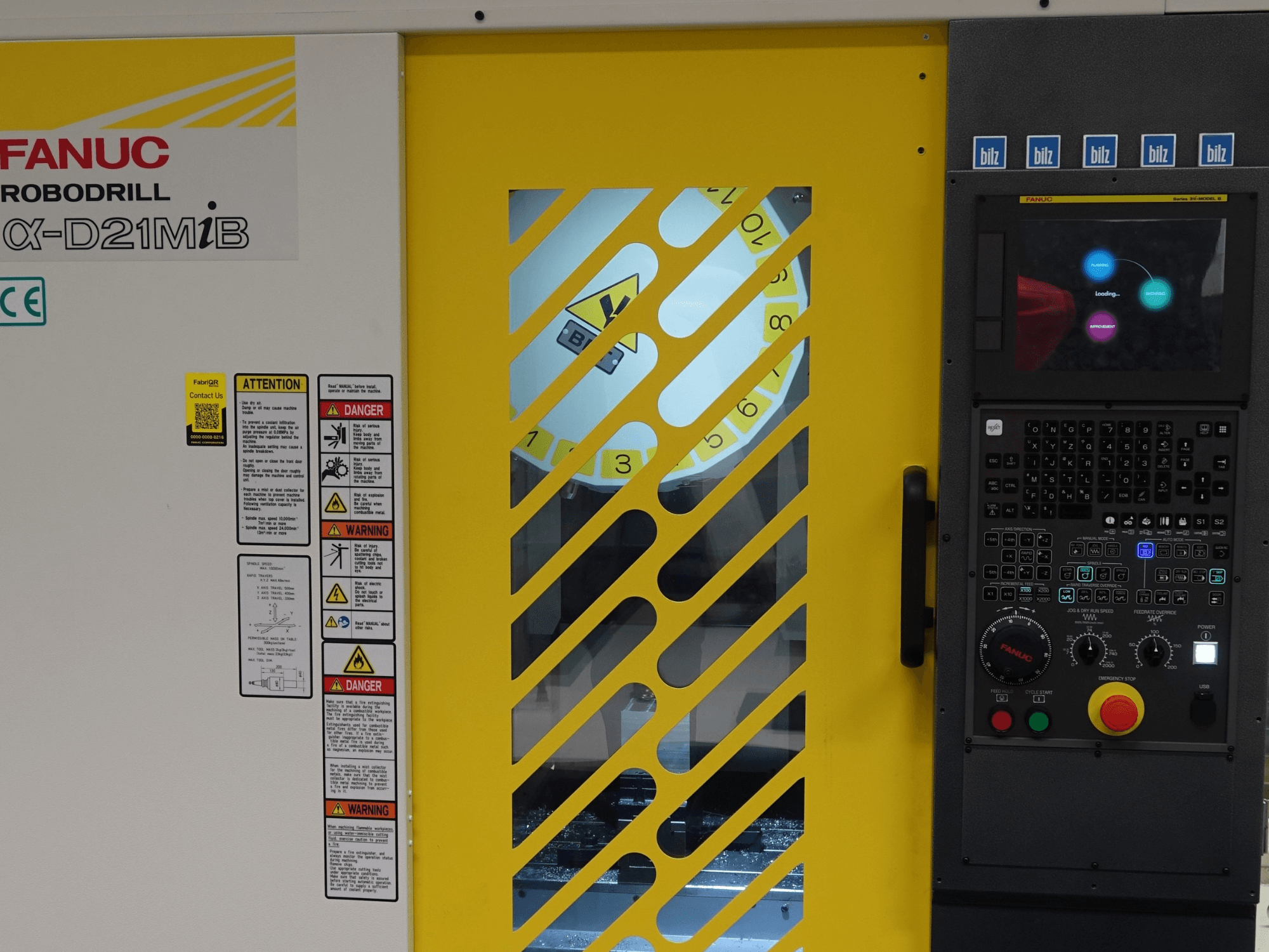 FANUC α-D21MiB -pystykoneistuskeskus edestä katsottuna; kuvassa näkyvät turvallisuusvaroitukset, ohjauspaneeli ja keltainen turvaovi.
