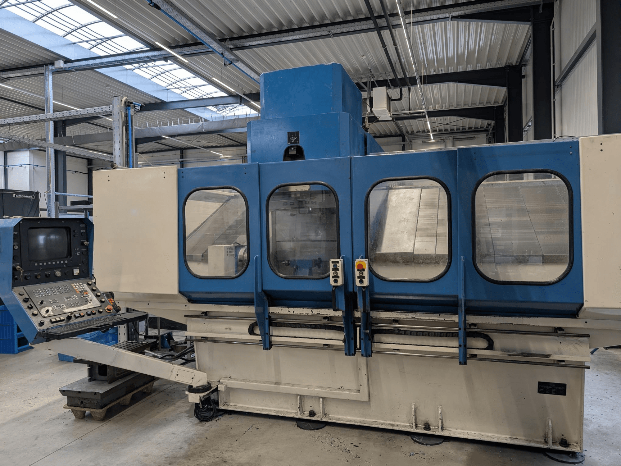Sininen teollinen CNC-kone, jossa on ohjauspaneeli, etukuva. Suuret lasi-ikkunat, näkyvät koneen osat ja painikkeet.
