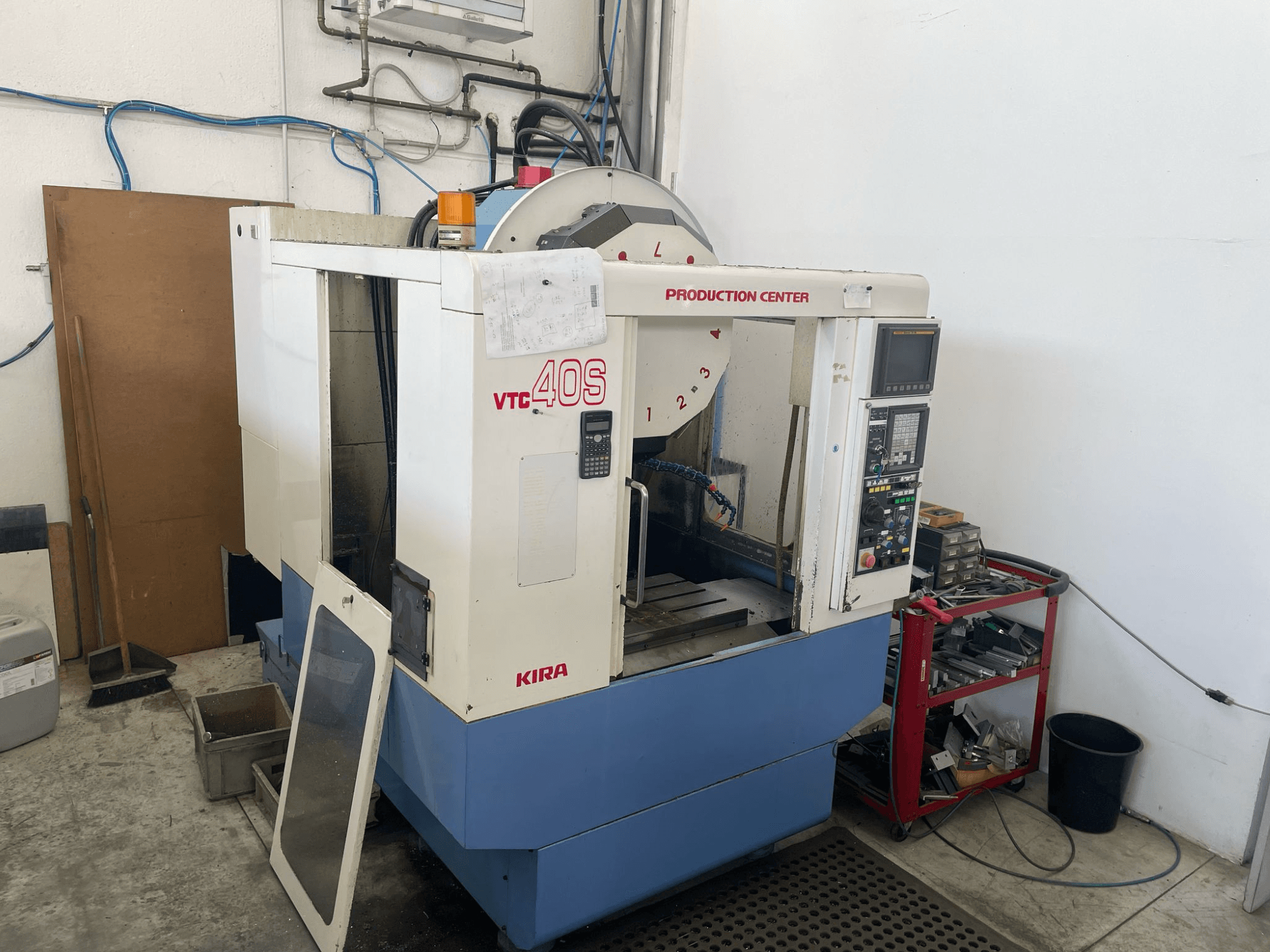 KIRA VTC 40S -työstökeskus sivukuvassa, jossa on ohjauspaneeli, avoin ovi ja työkalut varastokärryllä vieressä.