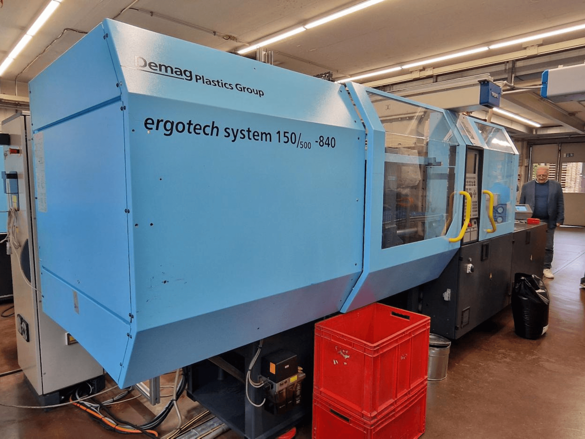 Demag Ergotech System 150/500-840 -ruiskuvalukone, sininen, sivukuva, taustalla laatikot ja käyttäjä.