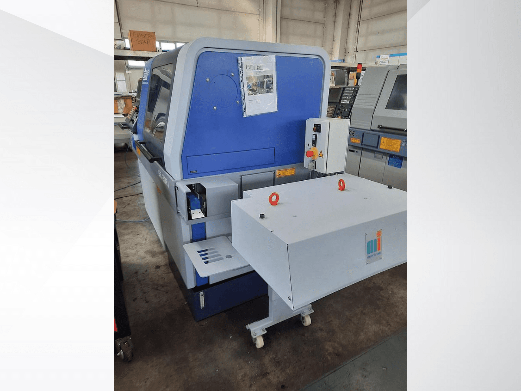 Sininen teollisuuskone valkoisella kuljettimella, jossa näkyy ohjauspaneeli ja siihen kiinnitetyt turvalaitteet, sivukuva.