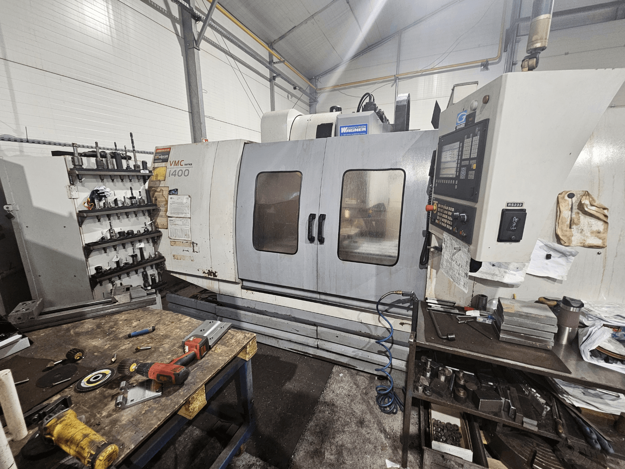 VMC 1400 CNC-jyrsinkone työpajassa, etukuva; työkaluja ja materiaaleja hajallaan työpöydällä lähellä.