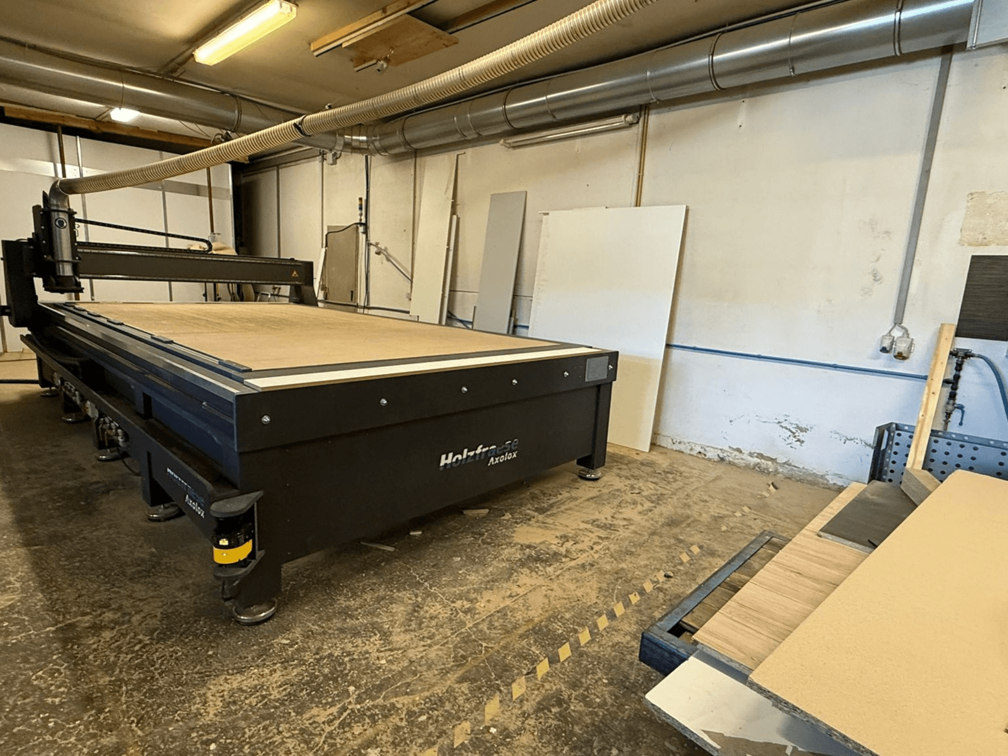 Holz-Her Axos 5 CNC-jyrsin työpajassa, etukuva, jossa näkyy työpöytä, pölynpoistojärjestelmä ja puulevyt lähellä.