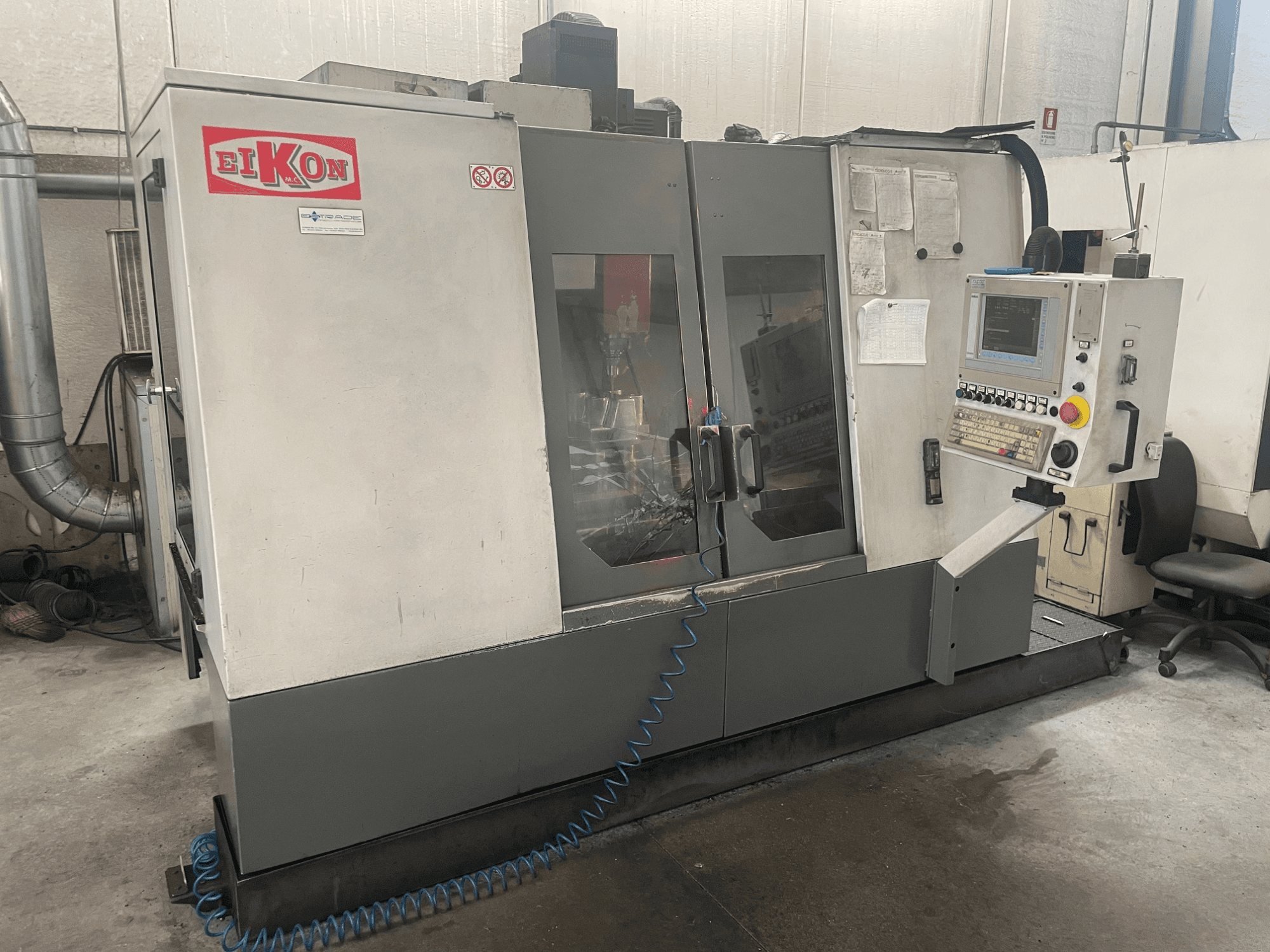 EIKON CNC-kone edestä katsottuna, jossa on ohjauspaneeli, suuret lasiovet ja sininen ilmaletku lattialla.