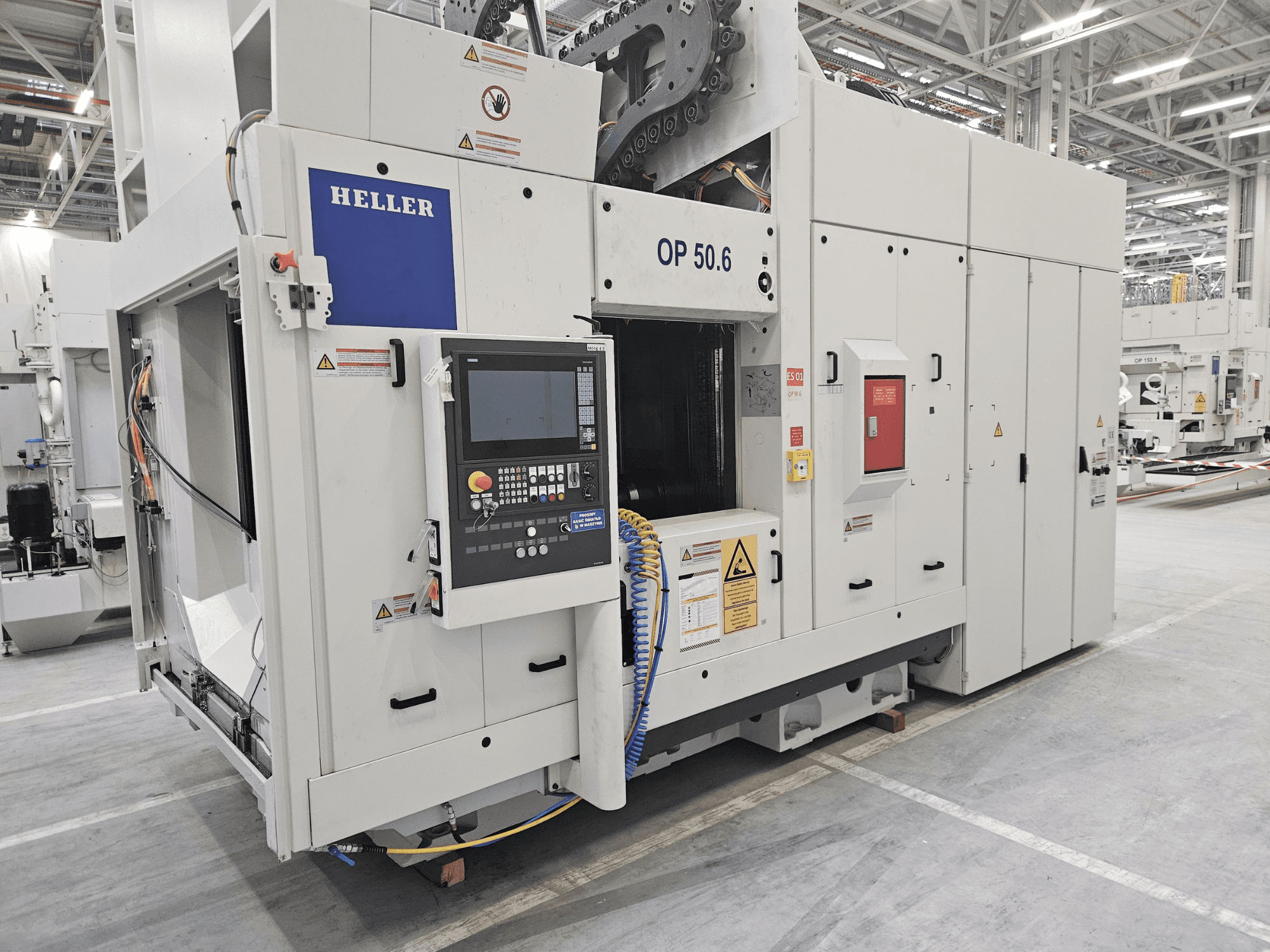 Heller OP 50.6 -työstökeskus, etukuva, jossa ohjauspaneeli, varoitustarrat ja letkut näkyvissä tehdasympäristössä.